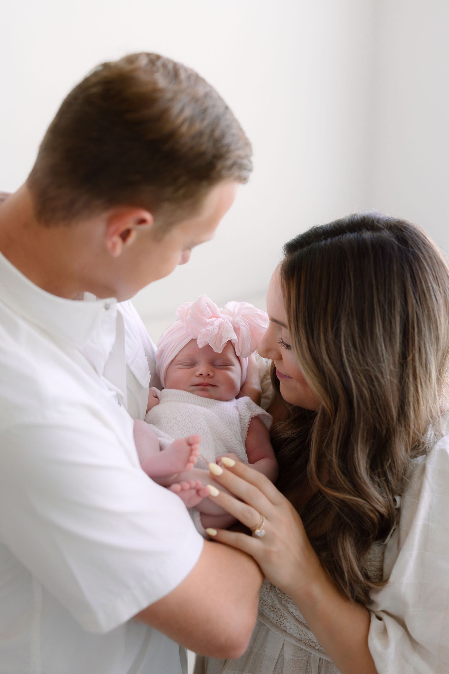 New parents lovingly gaze at their newborn baby girl in a pink bow headband during a professional newborn photo session.