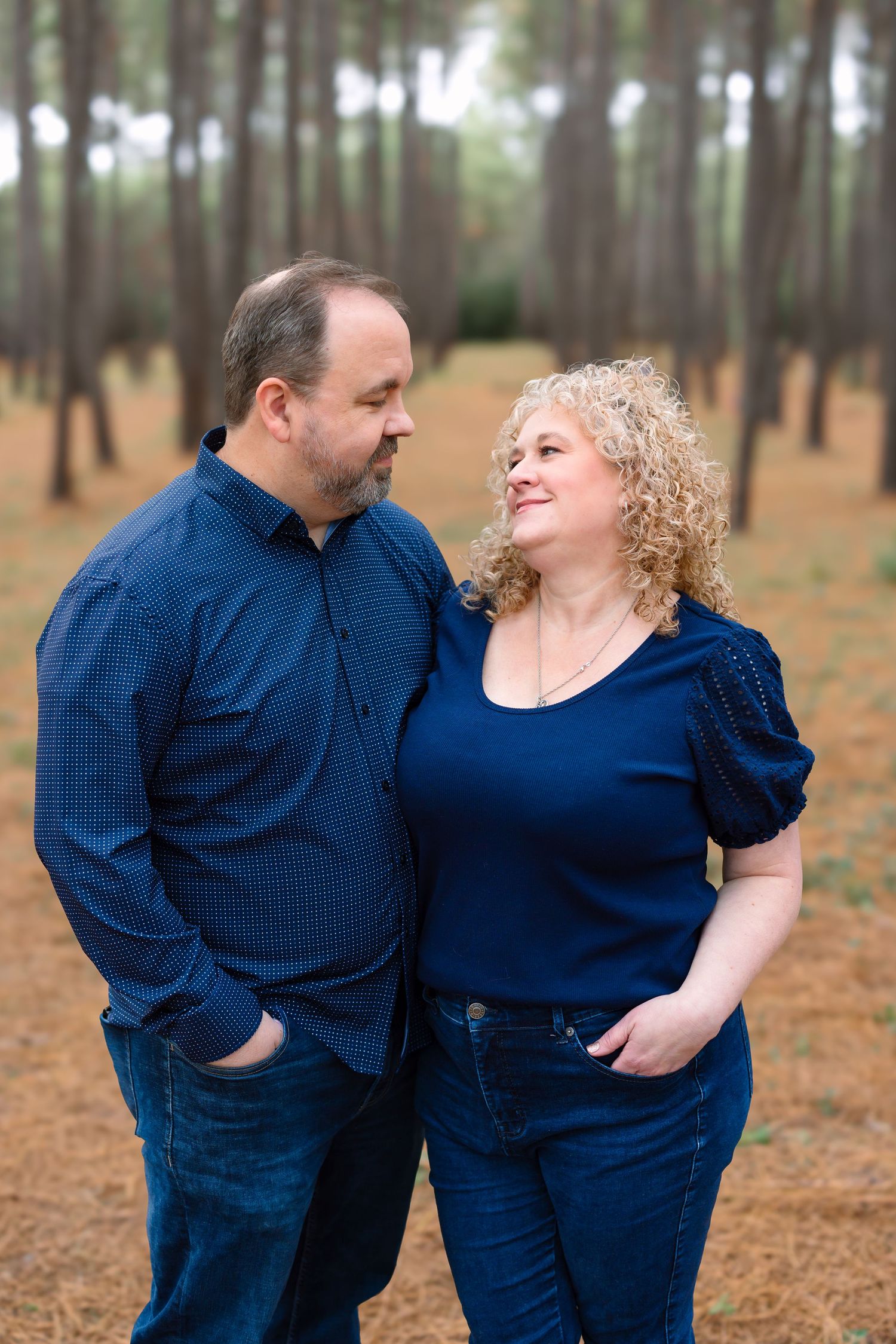 A couple in matching blue outfits share a tender moment among tall pine trees in a forest setting.