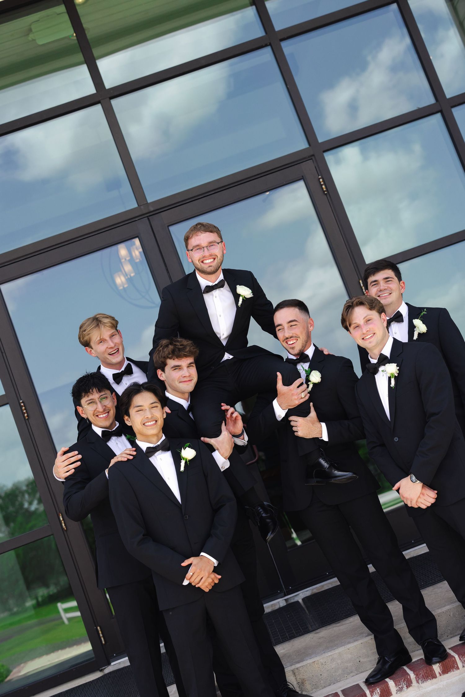 Groomsmen in black tuxedos pose together against modern glass building exterior.
