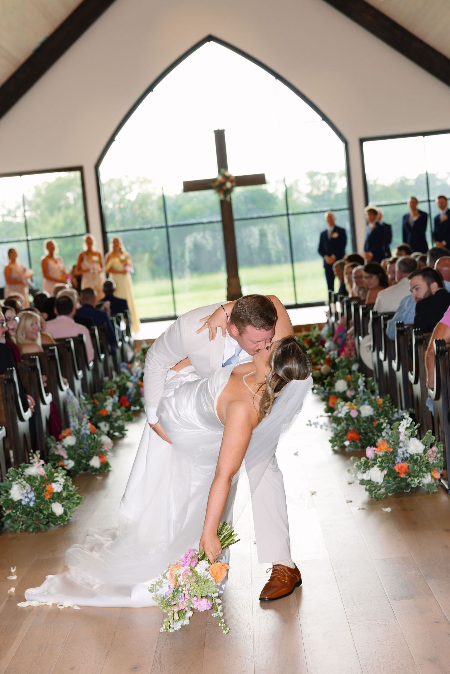 Bride and groom share a romantic dip kiss in a sunlit chapel aisle lined with colorful floral arrangements.