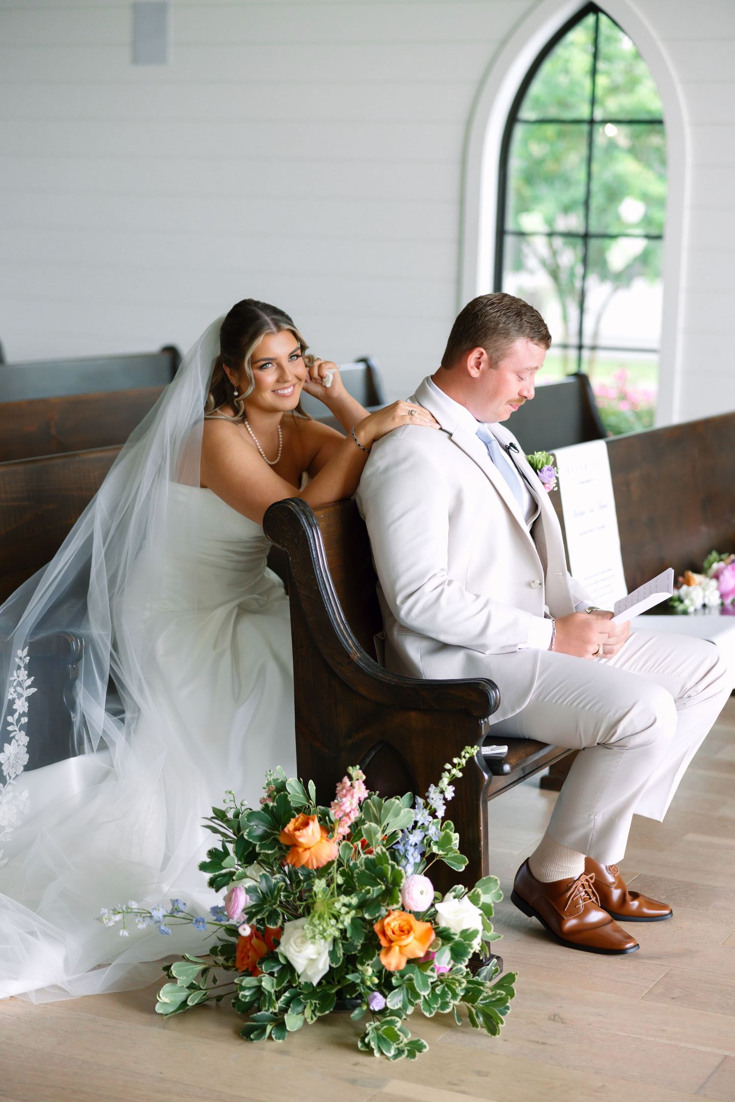 Bride and groom sit in church pew reading vows, with colorful bridal bouquet on floor in bright white chapel.