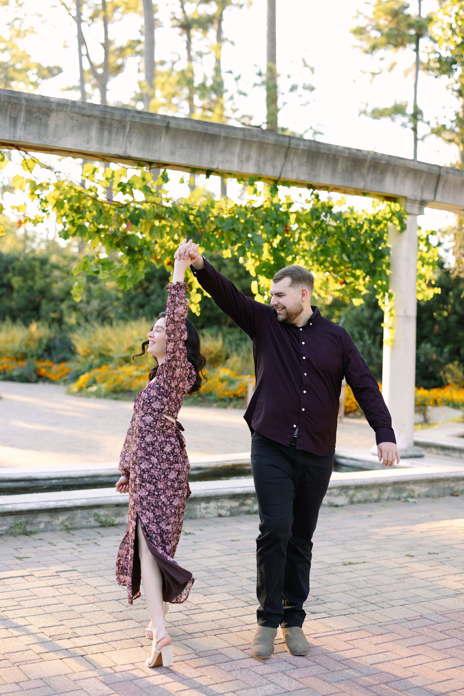 A couple dances joyfully outdoors, man in dark shirt spinning woman in floral dress on a brick patio surrounded by lush greenery.