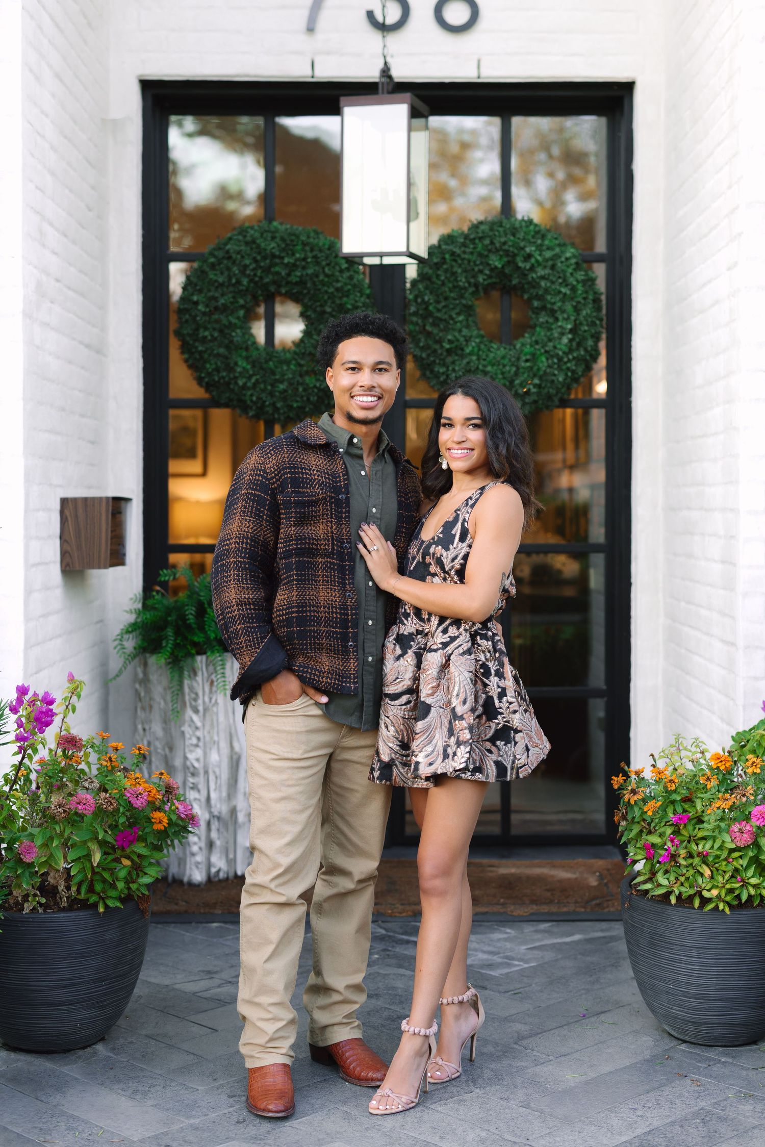 Smiling couple posing in front of a white brick building with green wreaths and colorful flowers at entrance 738.