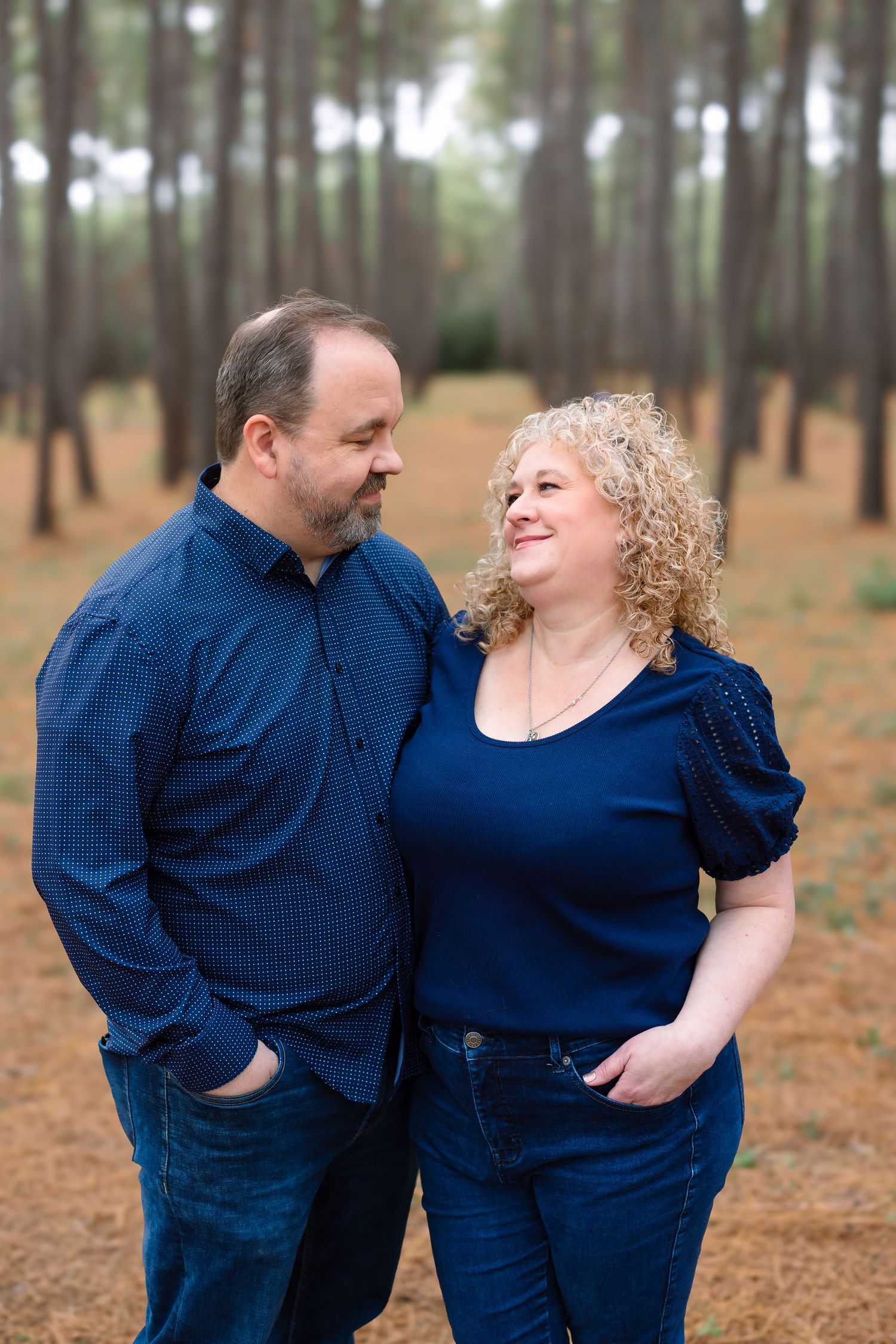 A couple in coordinating blue outfits share a warm smile while standing among tall pine trees.