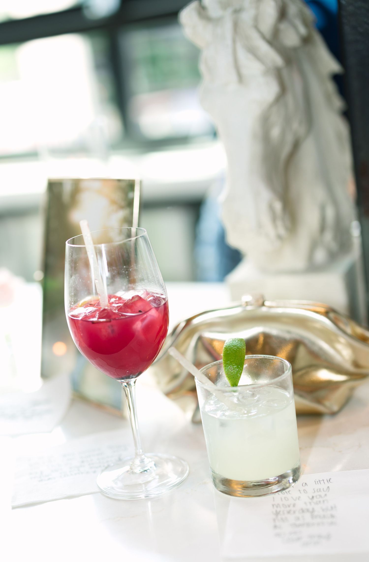 Two elegant cocktails on a white table, a red wine glass with crimson drink and a rocks glass with lime garnish.