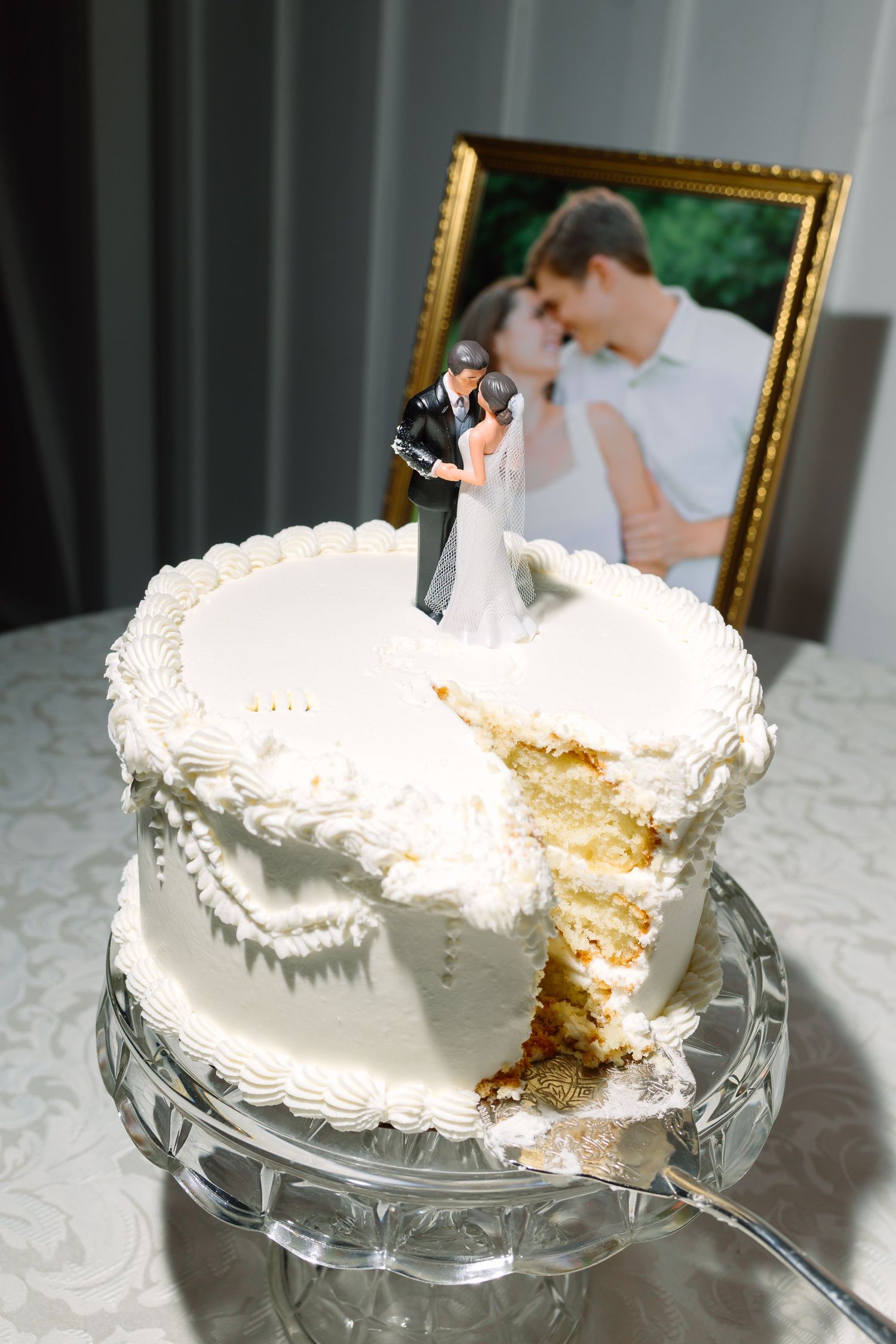 White wedding cake with bride and groom topper, partially sliced, displayed on crystal stand with couple photo in background.