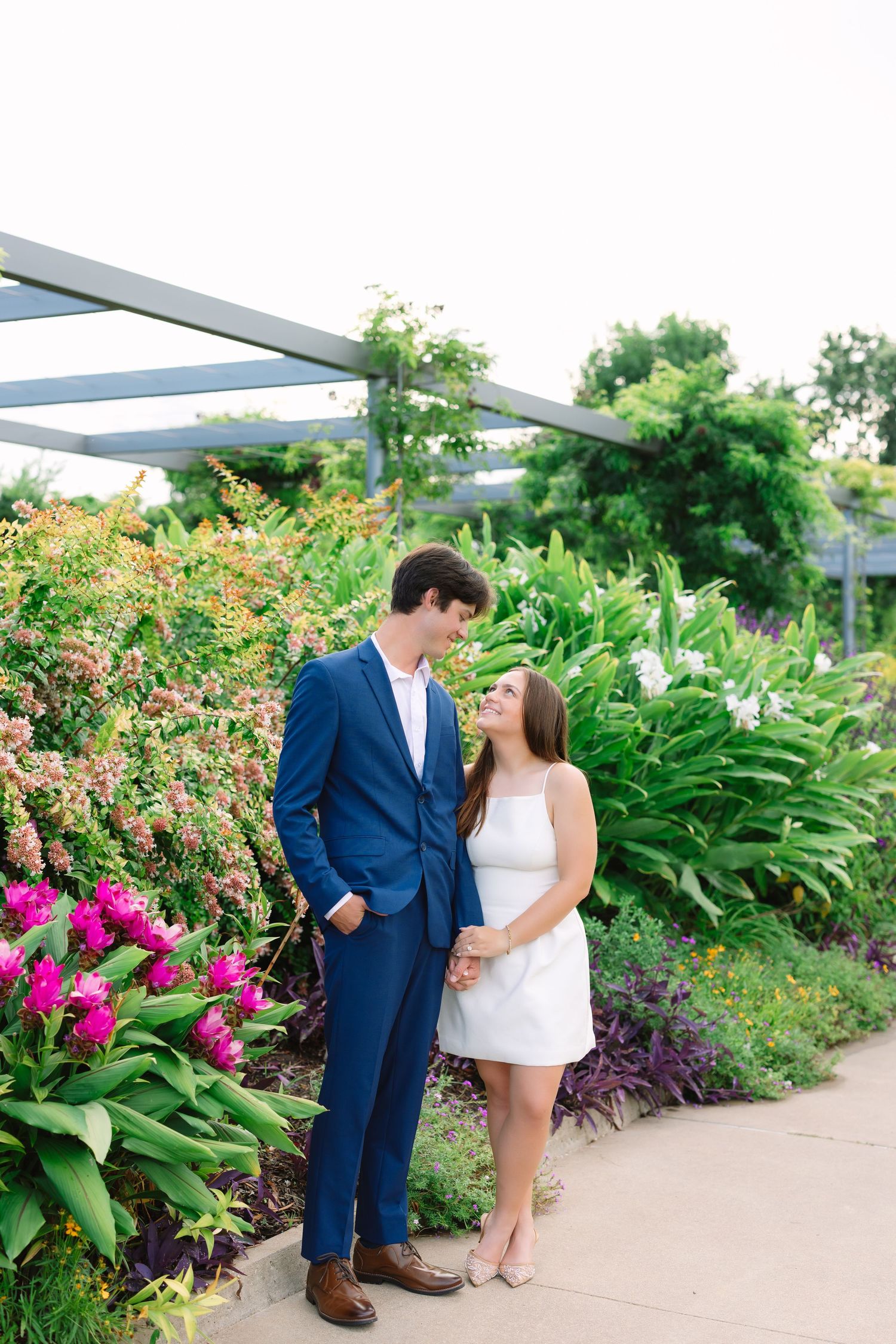 A couple poses playfully along a garden walkway surrounded by vibrant pink flowers and tropical plants.