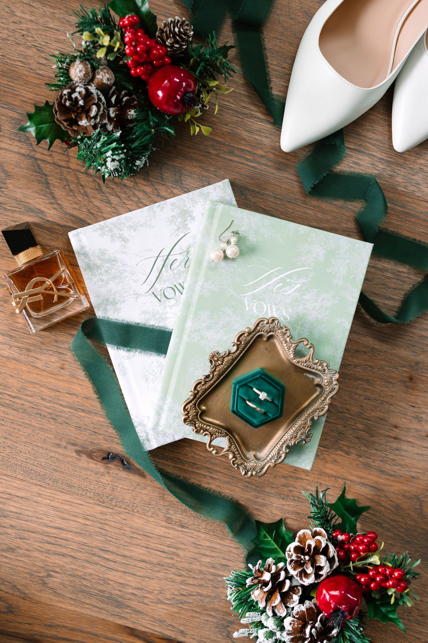 Green and white wedding stationery with rings, perfume, white heels, and Christmas pine cone decorations on wood.