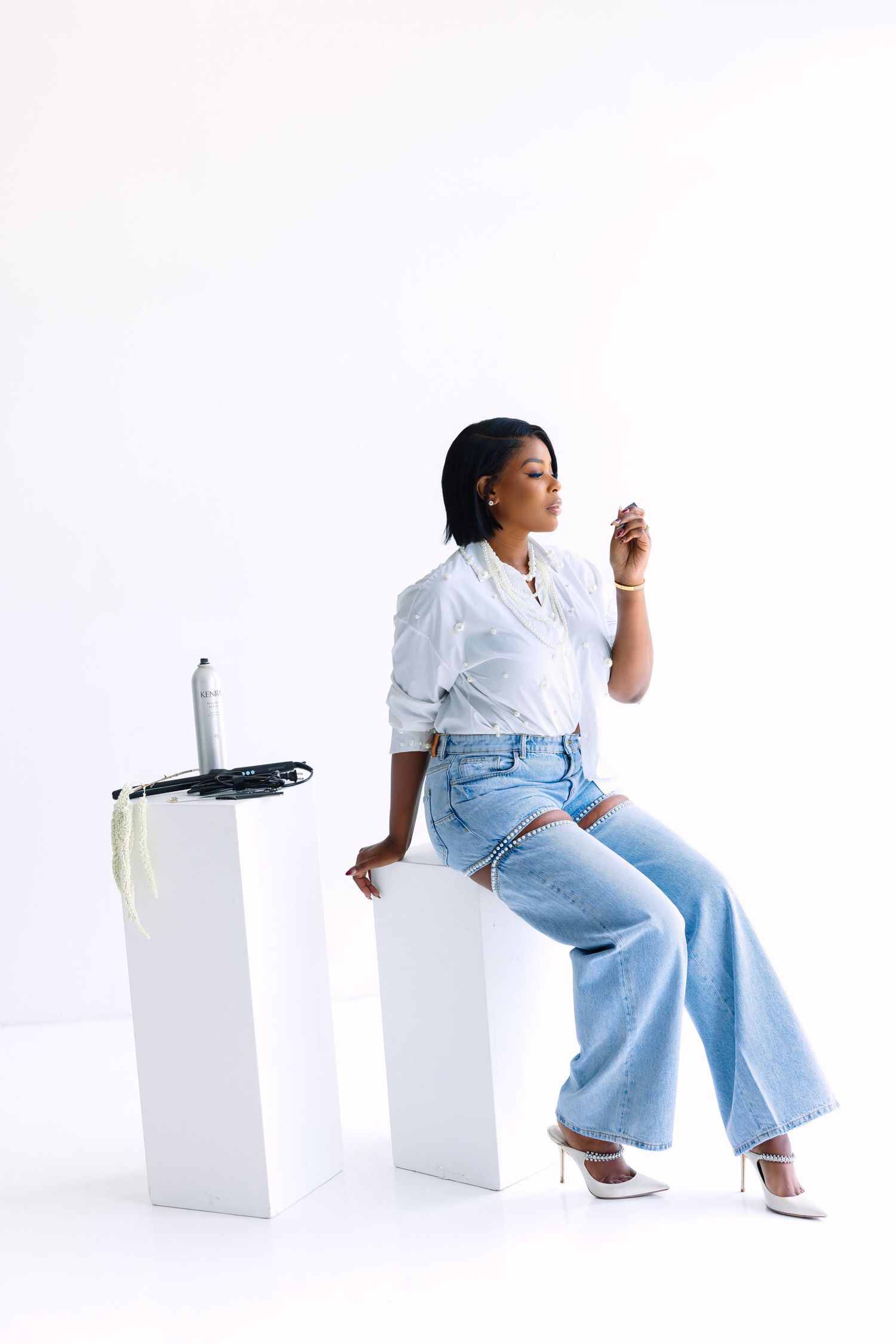 Woman in white shirt and ripped blue jeans sits on white cube, posing elegantly in white heels against a white background.