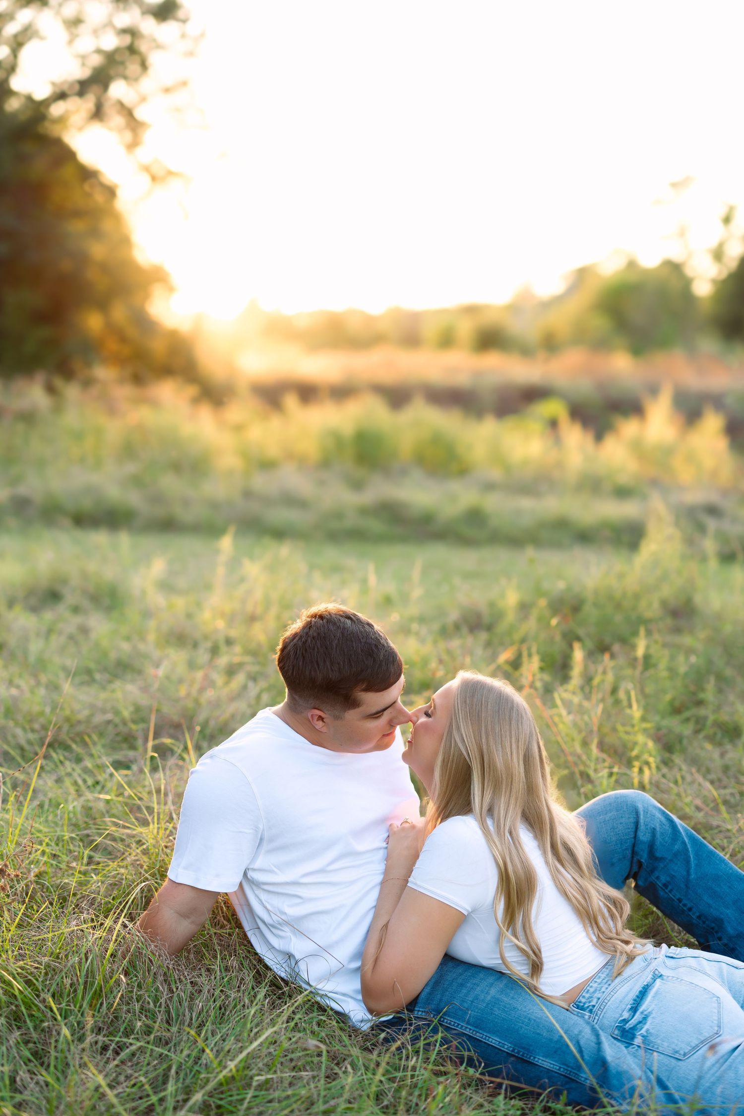 Romantic couple sitting in a grassy field at sunset, leaning in for a kiss in casual white tops and jeans.
