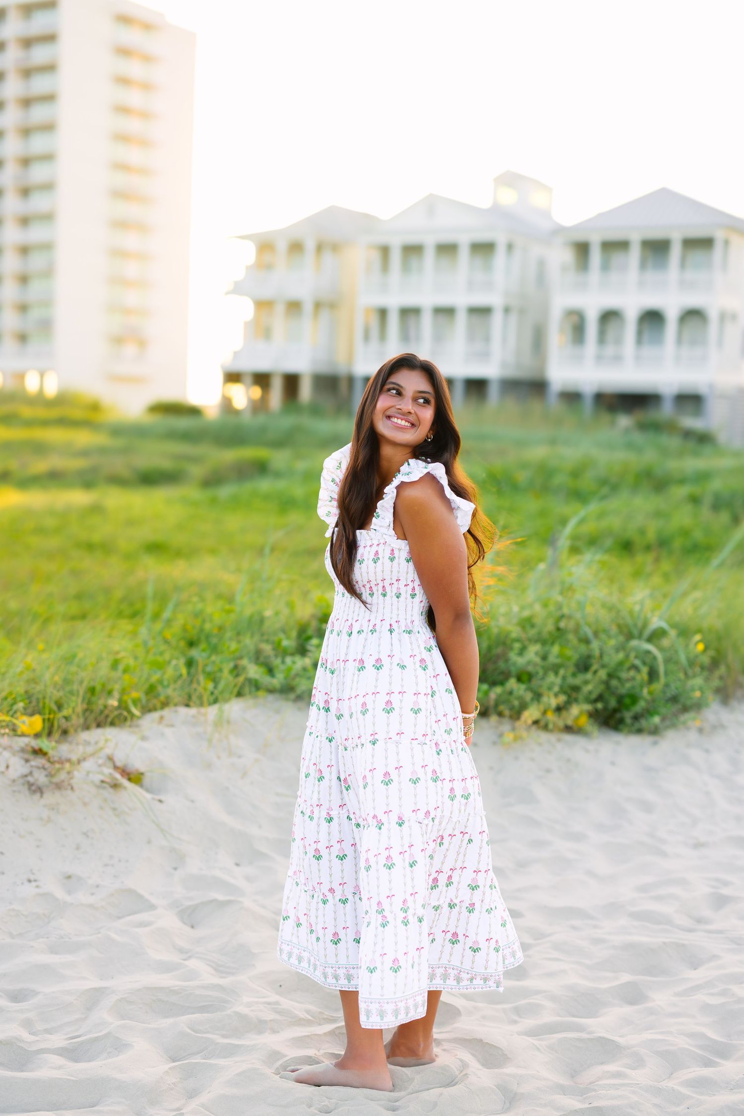 Young woman in white floral midi dress smiling on sandy beach with coastal homes in background.