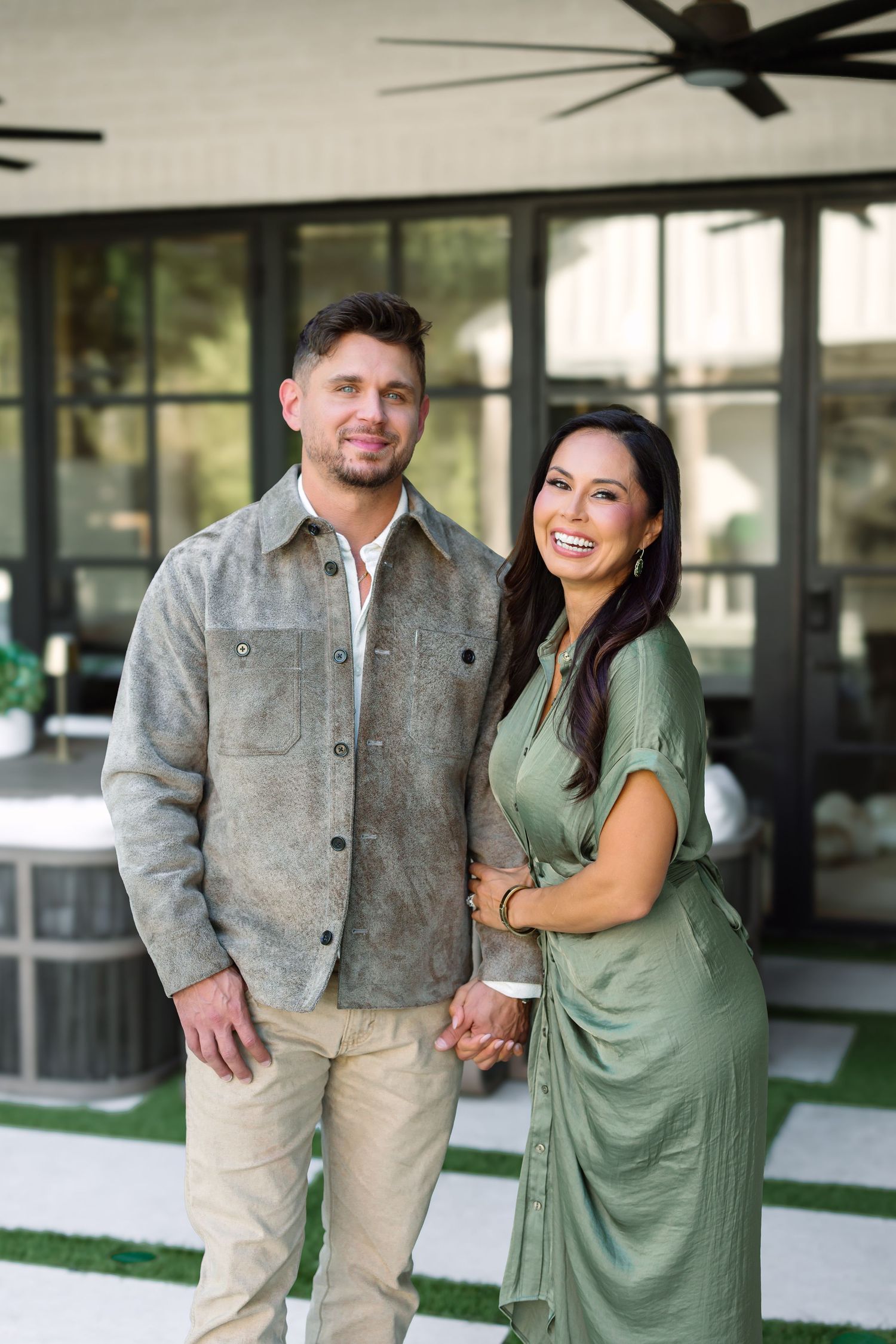 Smiling couple posing outdoors on a patio, man in gray jacket and khakis, woman in green dress.