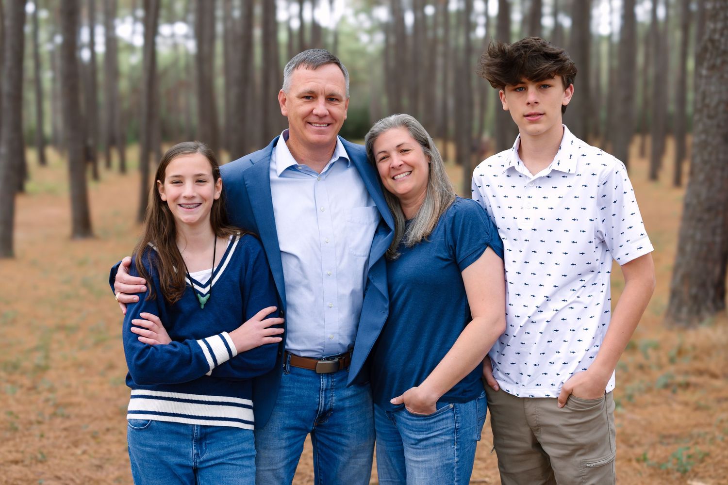 Family of four smiling together in a pine forest, wearing casual blue outfits for an outdoor portrait session.