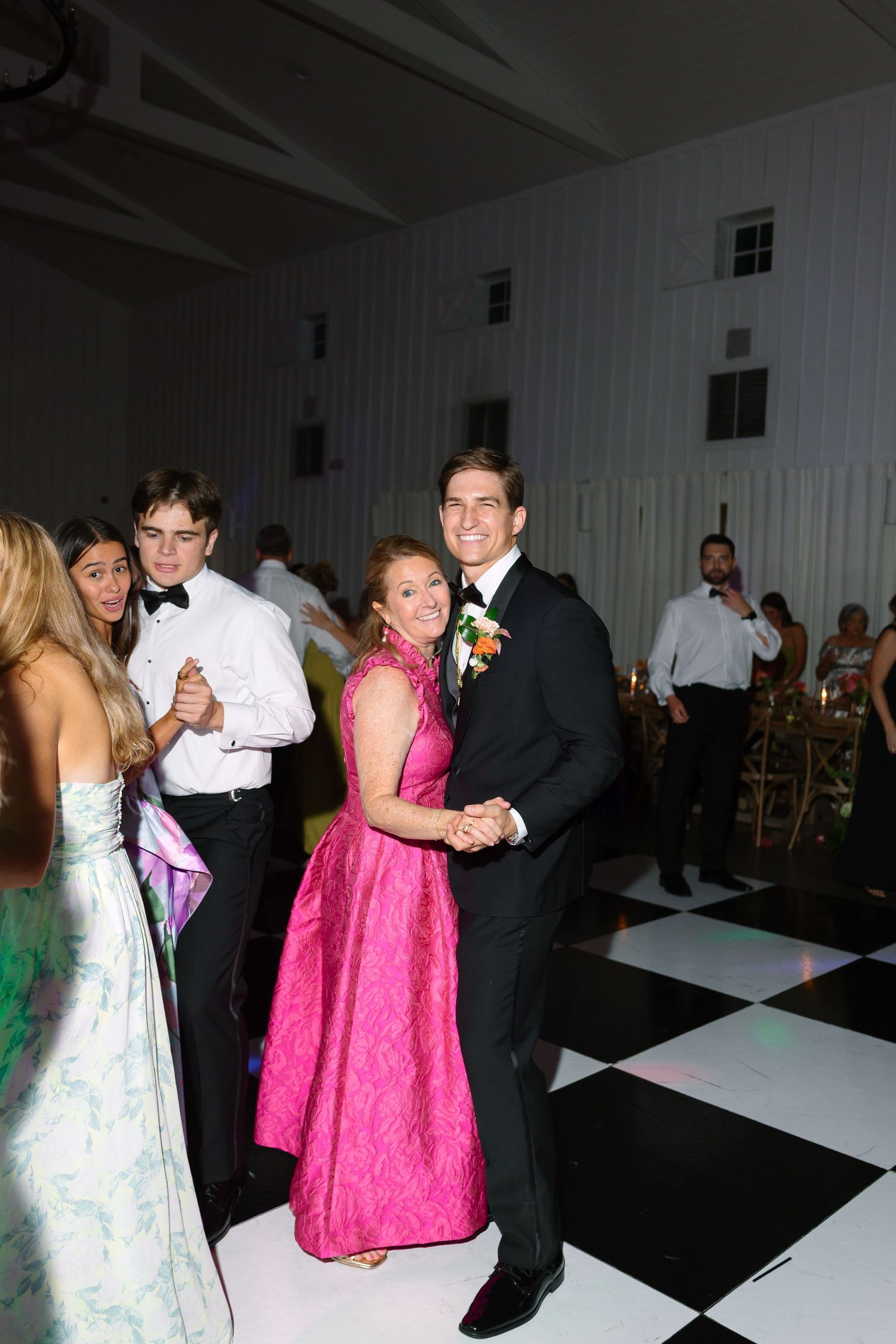 A couple dances elegantly on a black and white checkered floor at a formal event, woman in pink gown, man in black suit.