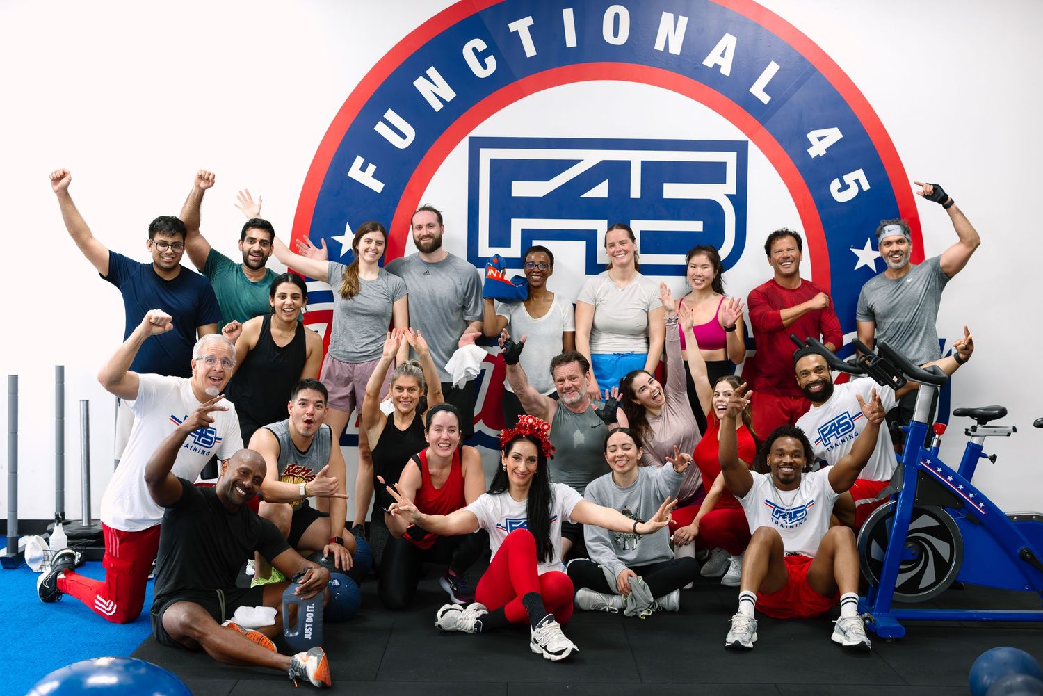Group of energetic F45 functional fitness class participants posing together in front of branded gym wall logo.