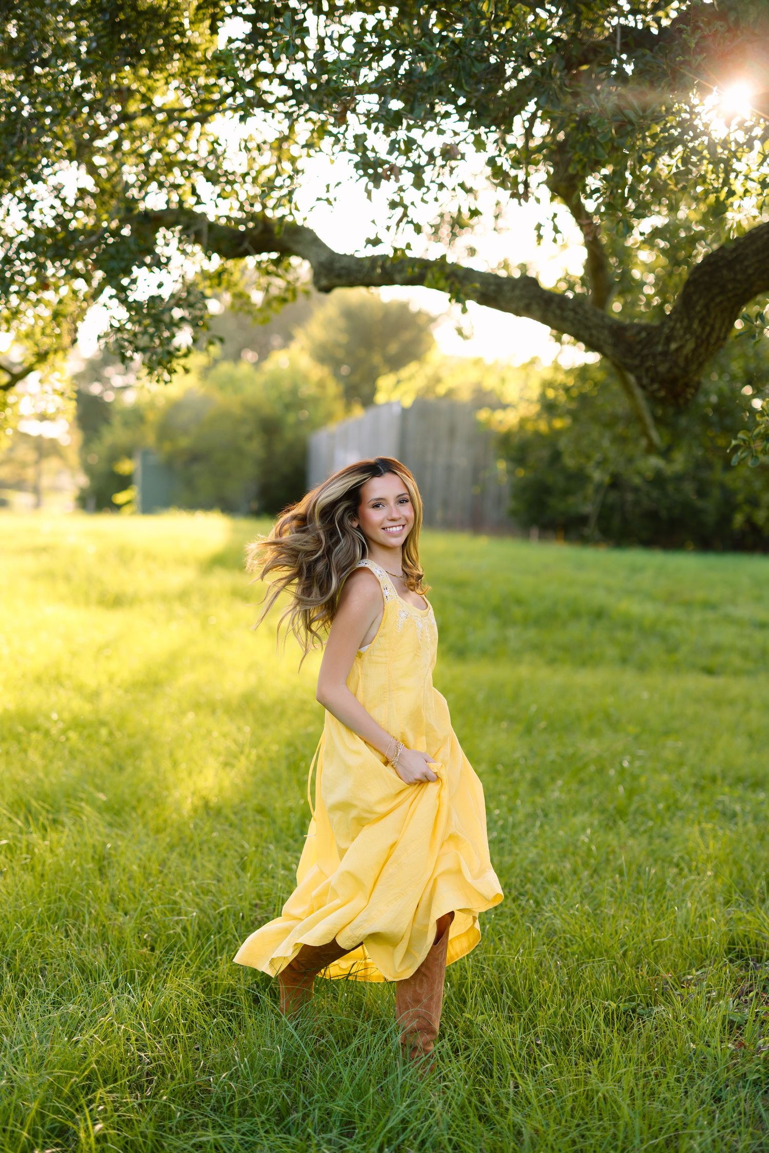 Young woman in yellow dress and brown boots twirling in sunlit green field under oak tree.
