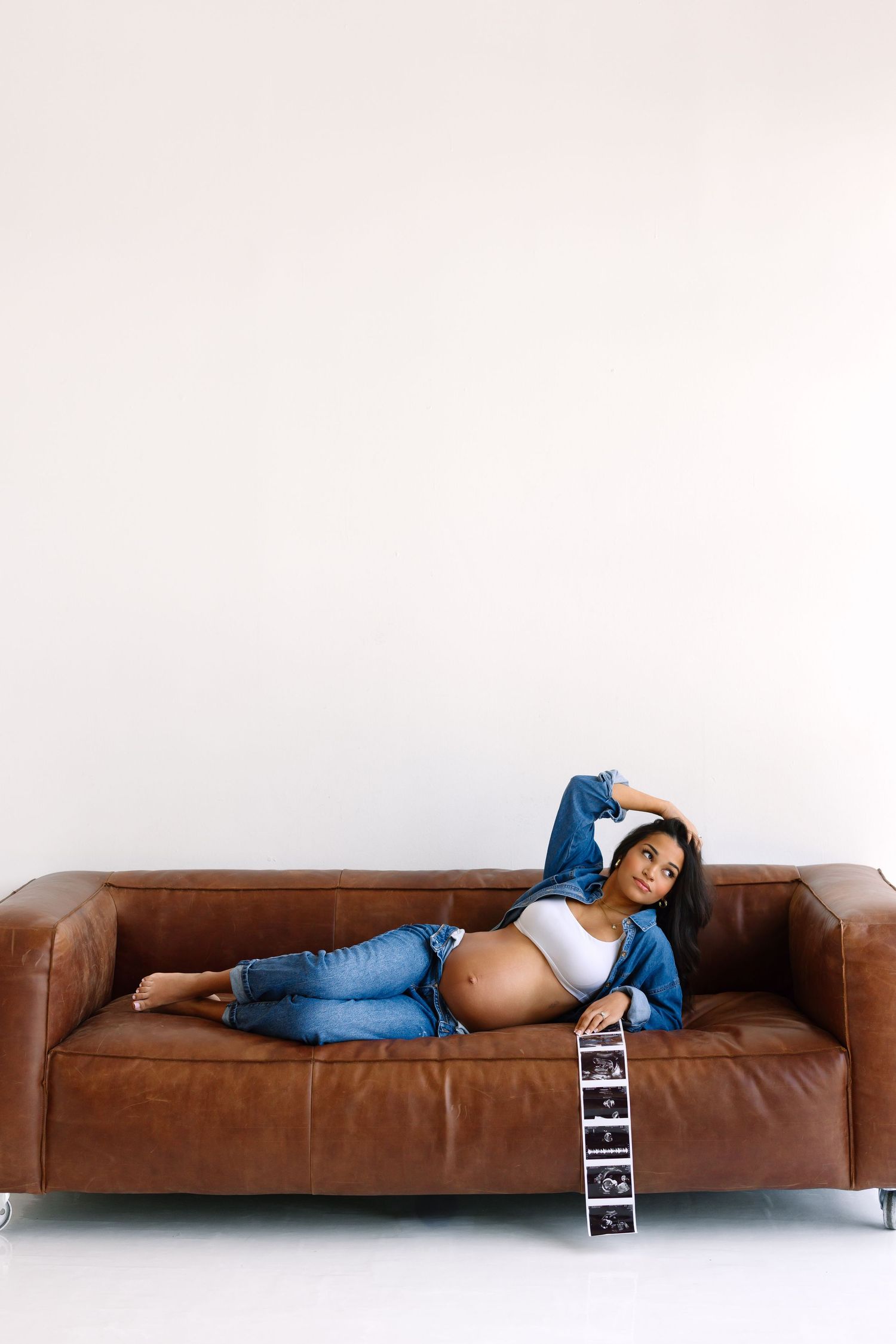 Pregnant woman in jeans lying on brown leather sofa holding ultrasound photos, showcasing baby bump.