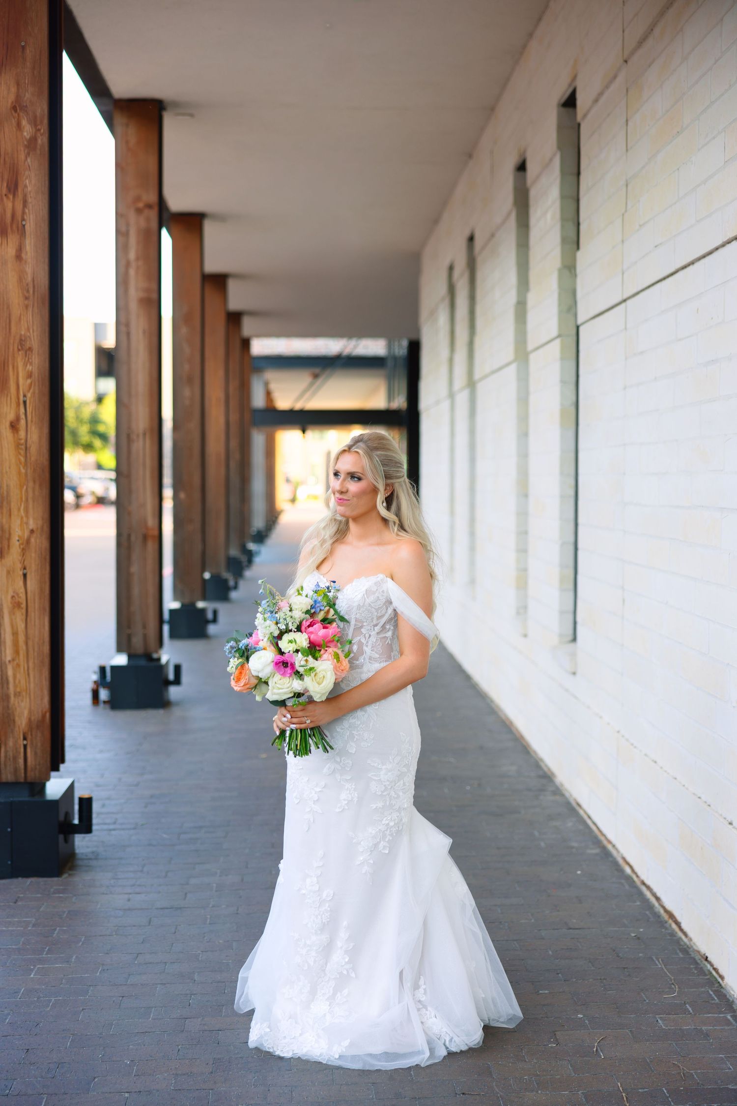 A blonde bride in an off-shoulder lace mermaid gown holds a colorful bouquet while posing outdoors near a modern building.
