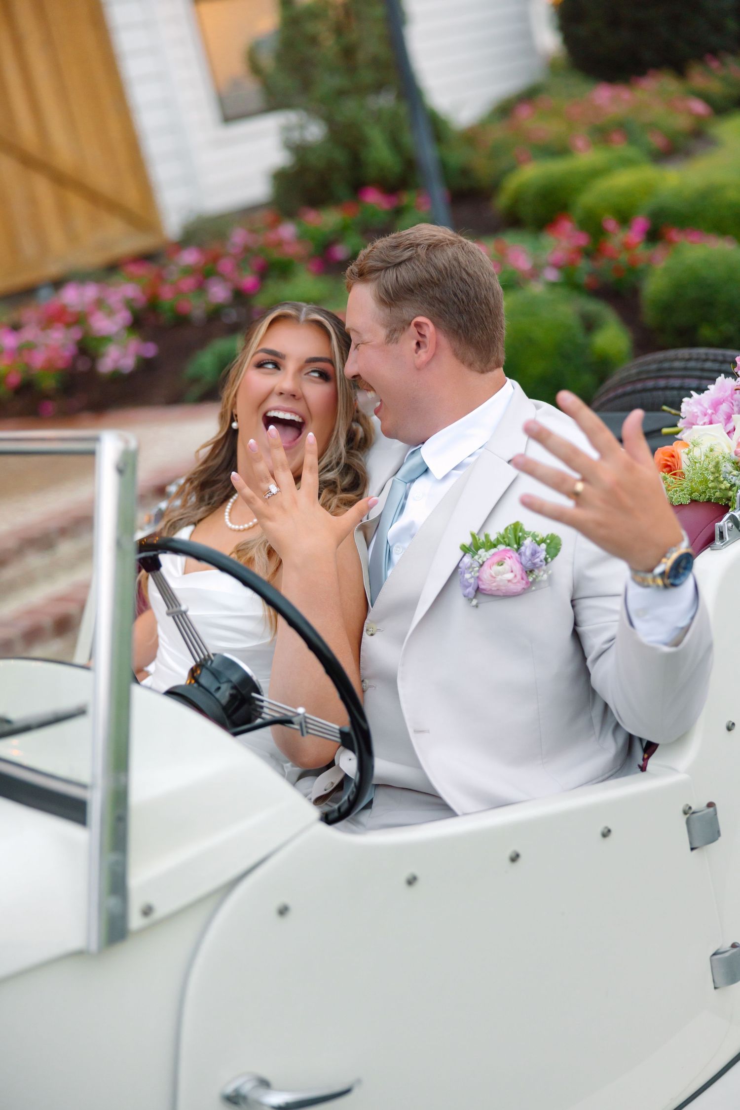 Happy newlyweds in a vintage white convertible car, bride showing ring, groom holding bouquet.