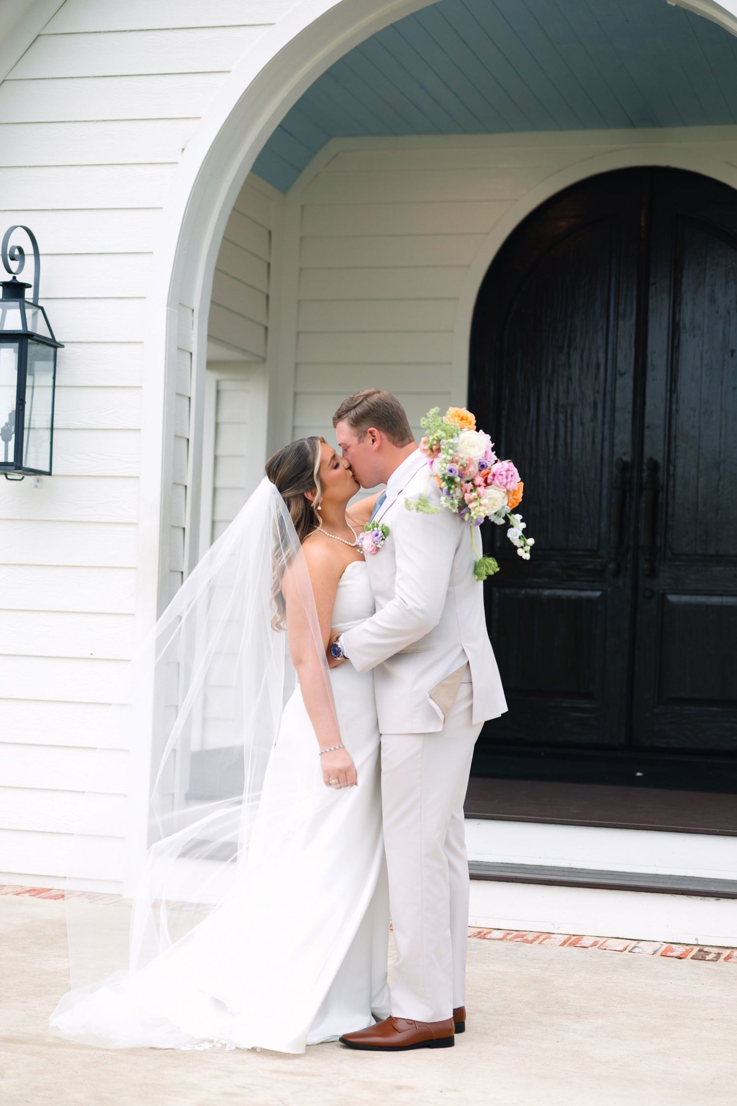 Bride and groom share a romantic kiss in front of a white chapel with a colorful bridal bouquet.