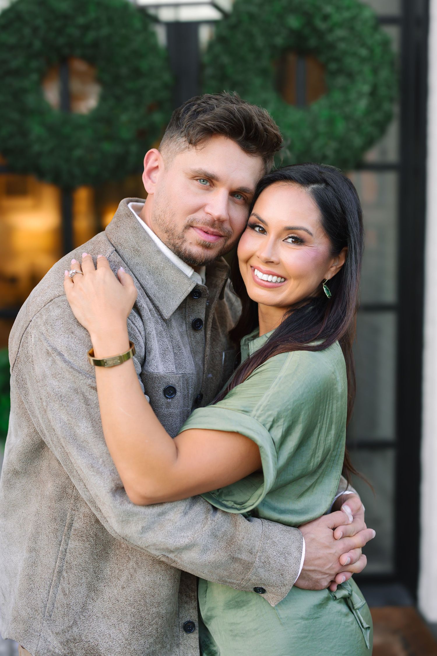 A smiling couple embraces warmly outdoors, wearing green and grey casual outfits near a wreath.