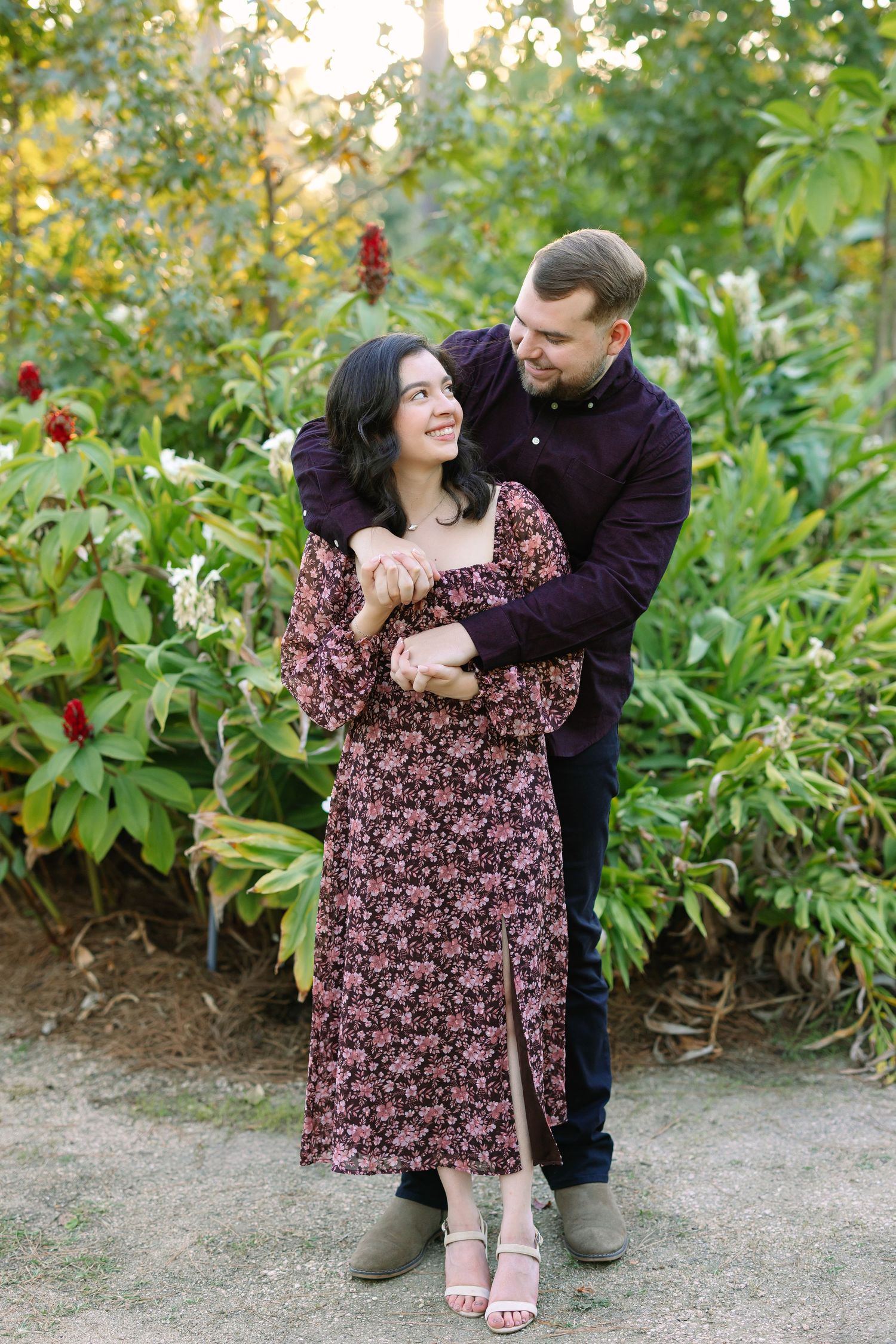 Couple embracing in a lush garden, woman in floral dress smiling up at man in dark purple shirt.