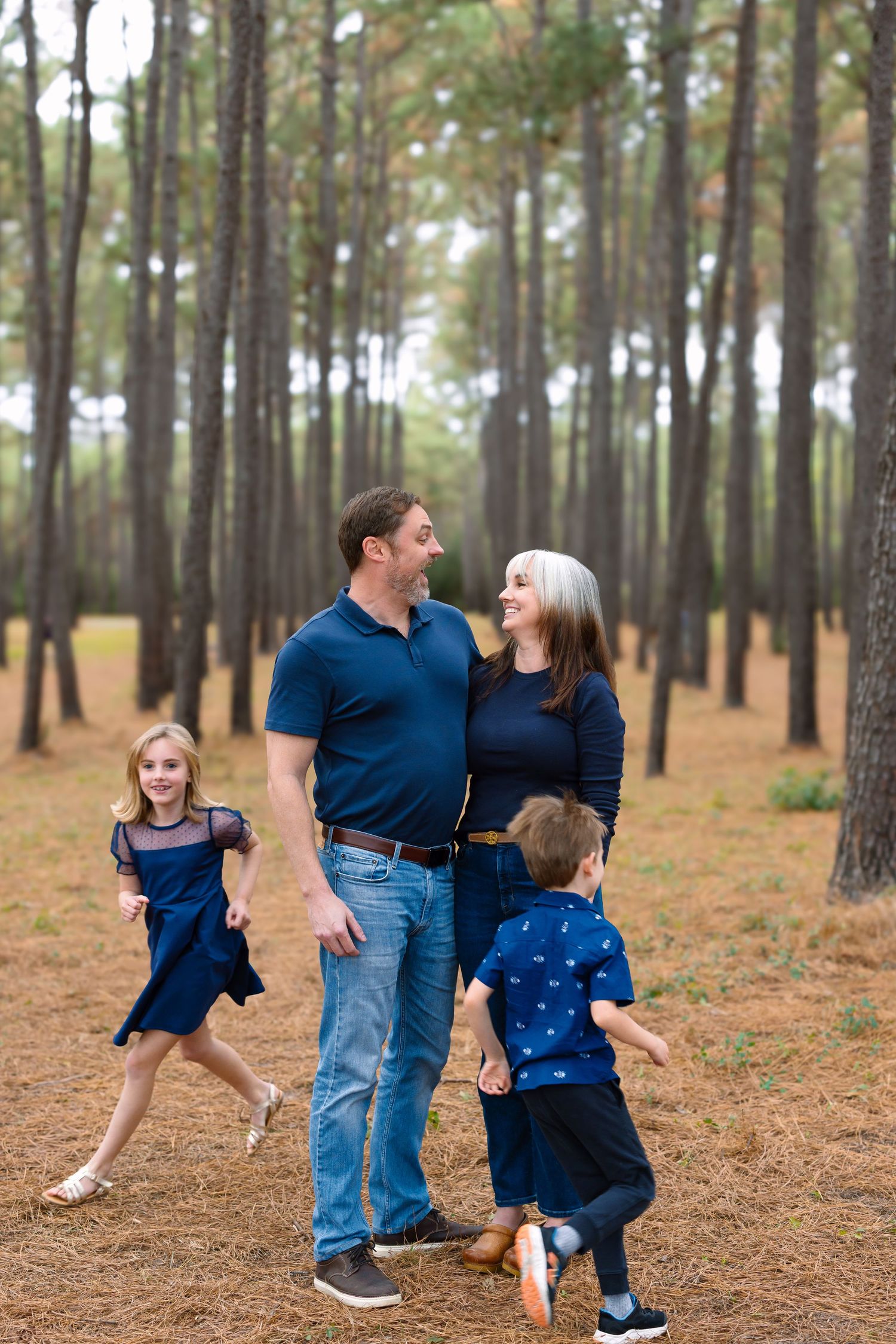 Family of four in navy blue outfits walking and laughing together in a pine forest.