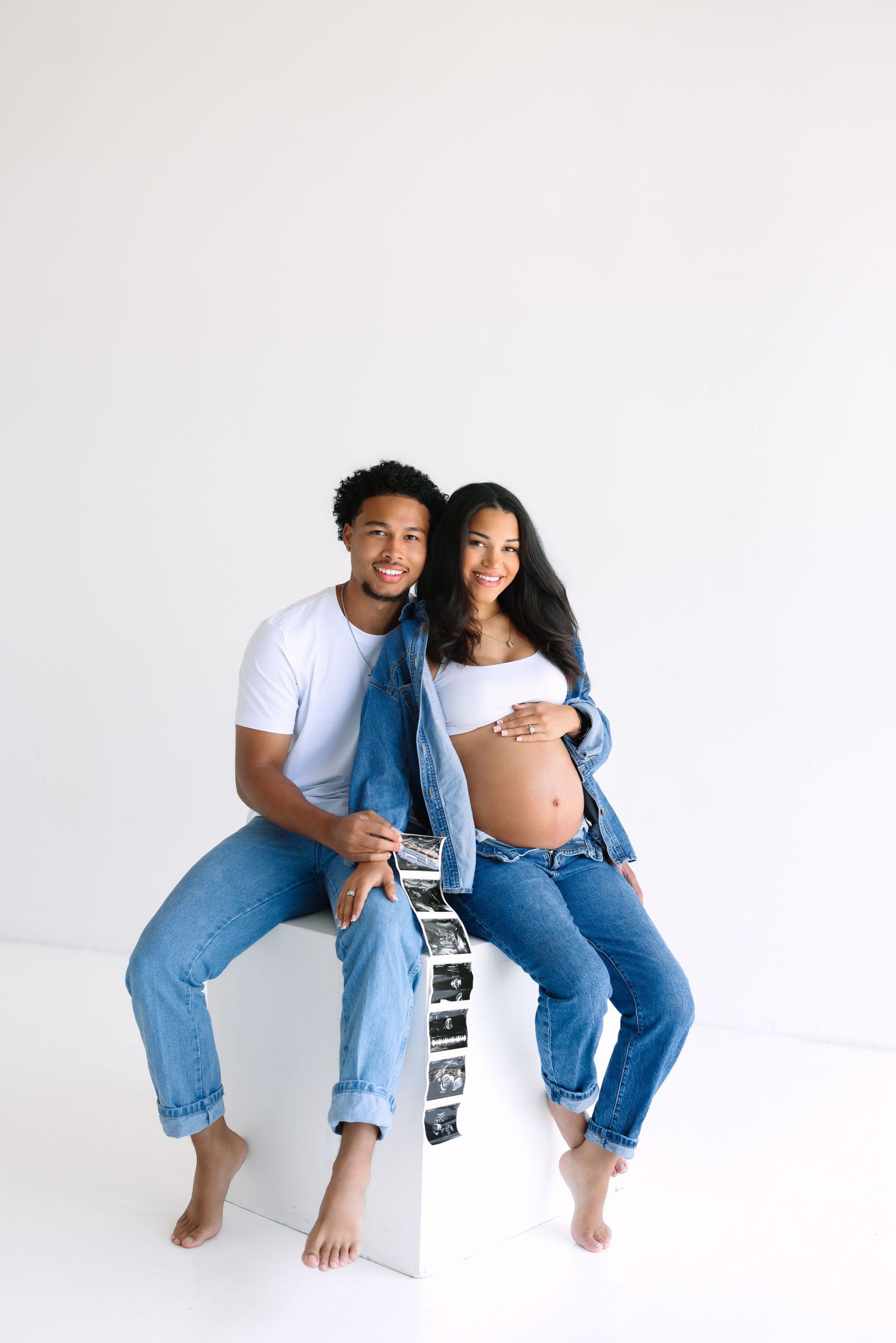Happy expecting couple in denim outfits sitting together, woman cradling baby bump, holding ultrasound photos on white background.