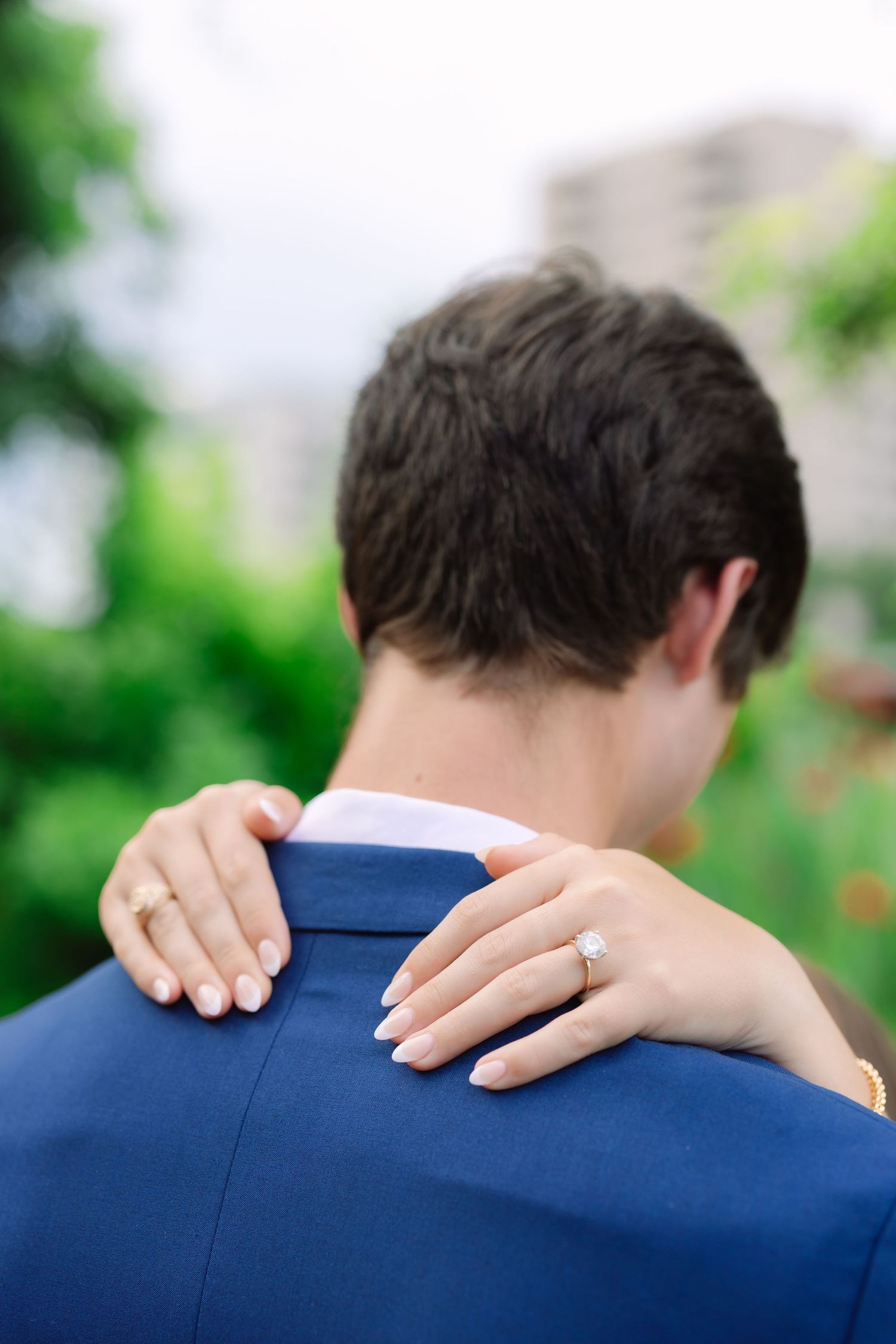 Close-up shot of hands resting on a navy blue suit jacket showing an engagement ring.