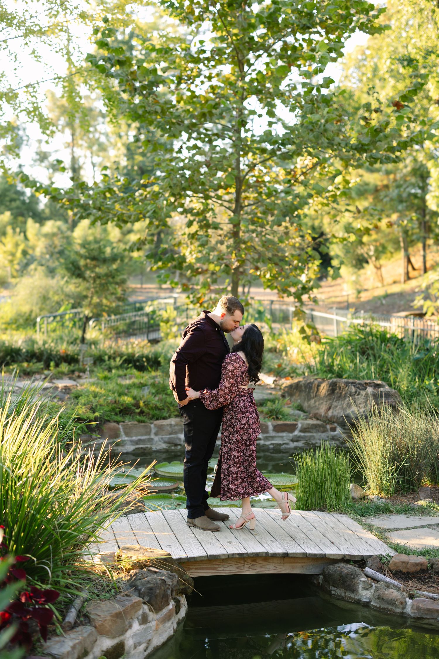 Couple sharing a romantic kiss on a wooden bridge over a pond in a lush green garden setting.