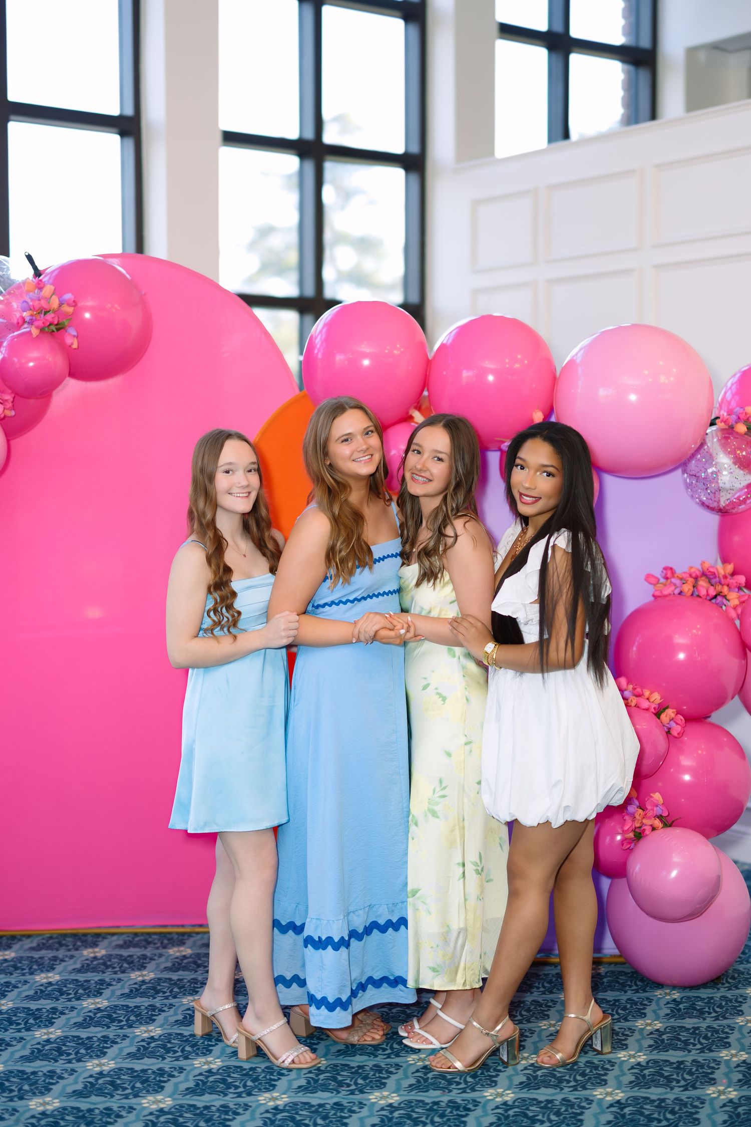 Four young women in colorful dresses pose together in front of a vibrant pink and orange balloon backdrop.