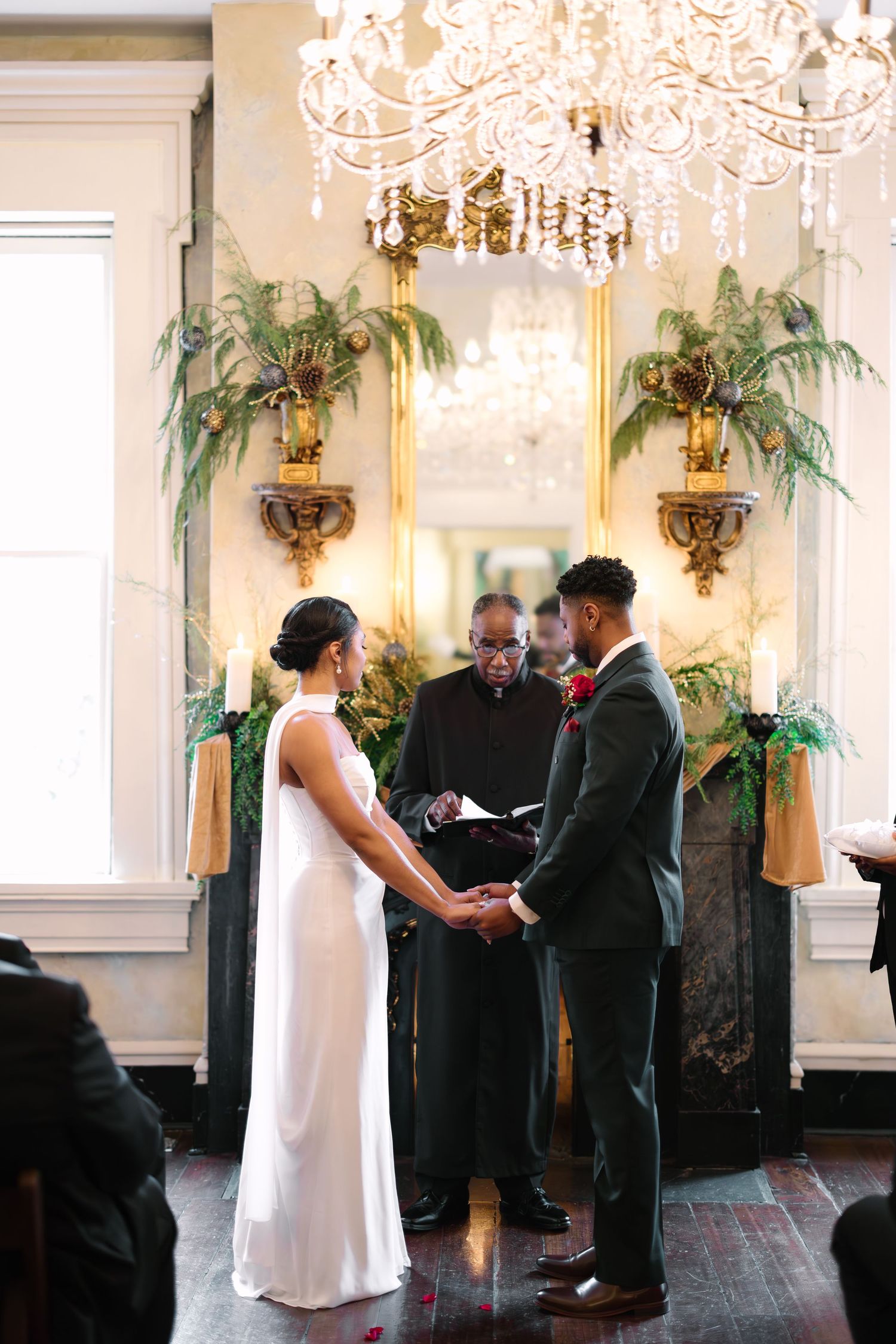 Couple exchanges vows at elegant indoor wedding ceremony with crystal chandelier and lush fern decor.