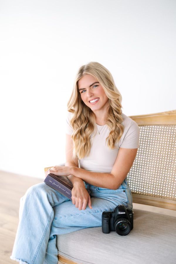 Woman in white t-shirt and jeans sits casually on rattan chair with camera nearby.