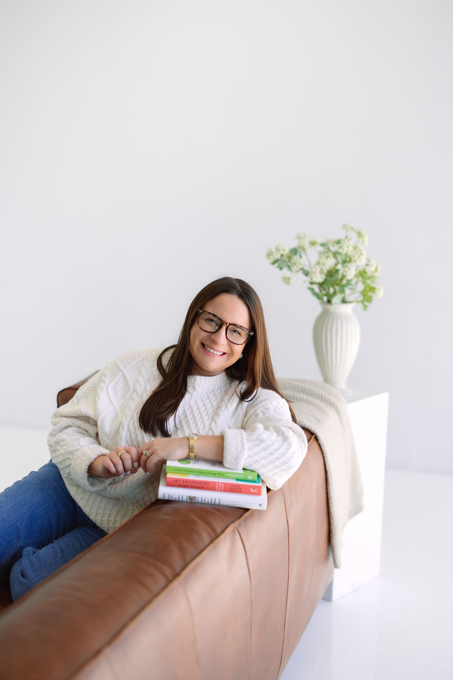 Smiling woman in white knit sweater and glasses relaxing on leather sofa with stacked books and flower vase.