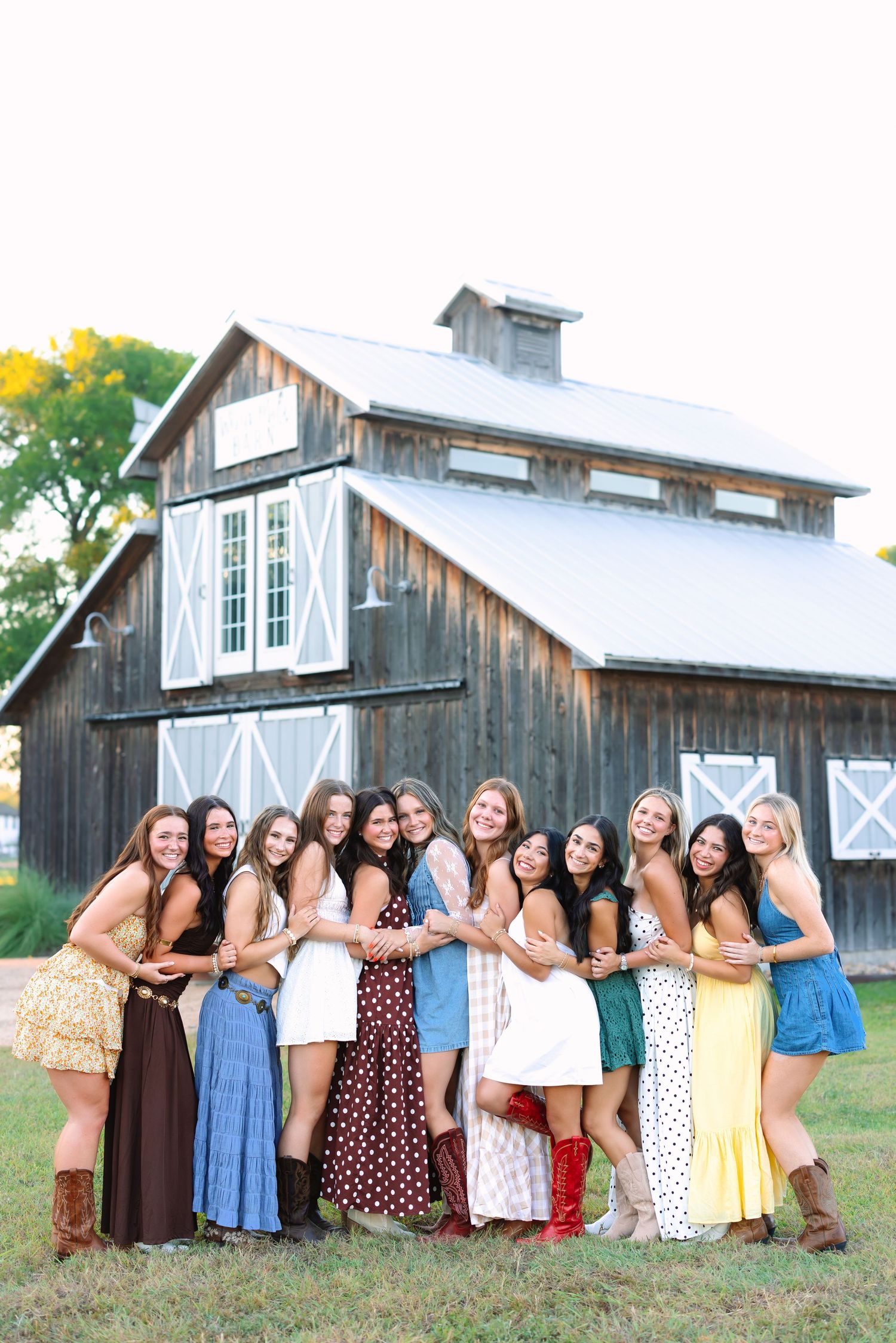 Friends in colorful summer dresses huddle close for a group photo by a barn.