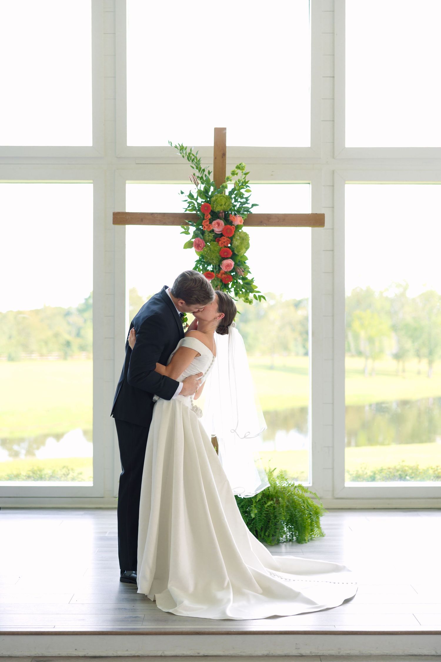 Bride and groom share first kiss under floral cross altar by large window at elegant indoor wedding venue.