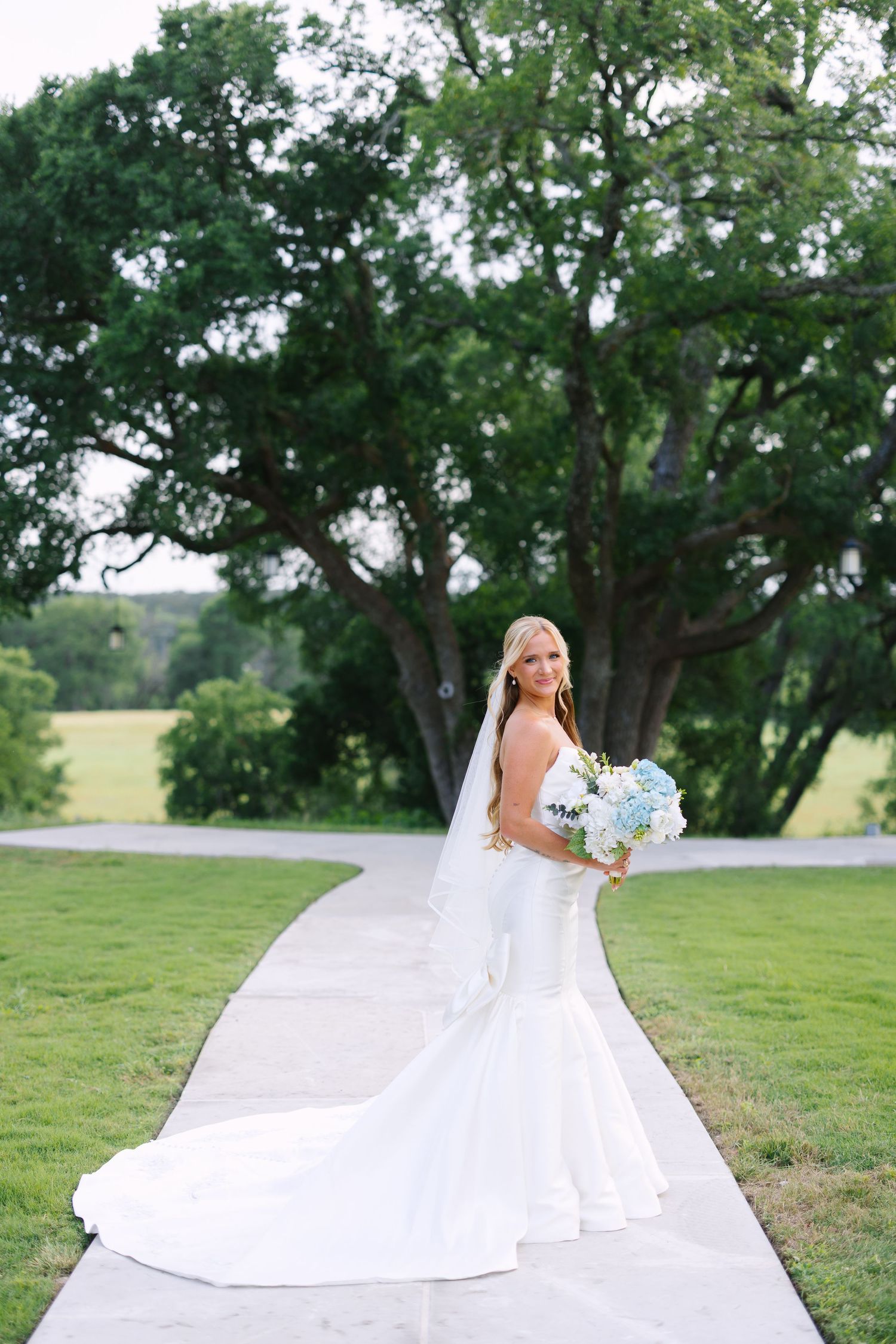A bride in a white ballgown poses outdoors with trees and rolling hills in the background.