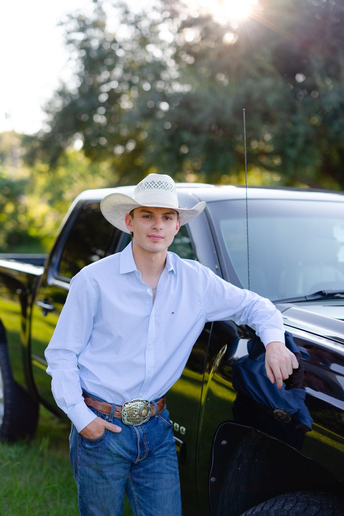 A person in a white cowboy hat and shirt leans against a black car in a sunlit outdoor setting.