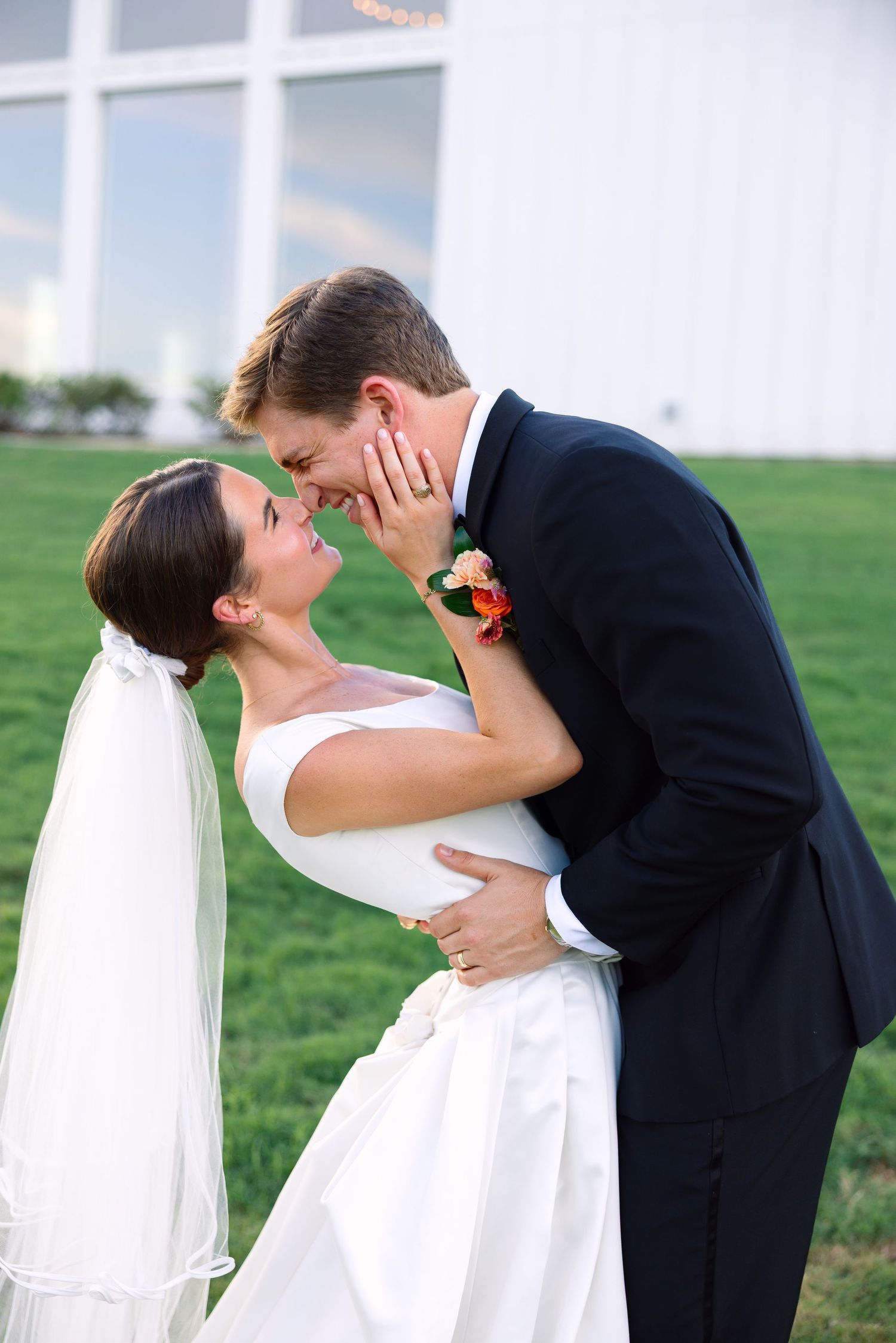 Bride and groom share a romantic moment outdoors, nearly kissing in wedding attire on green grass.