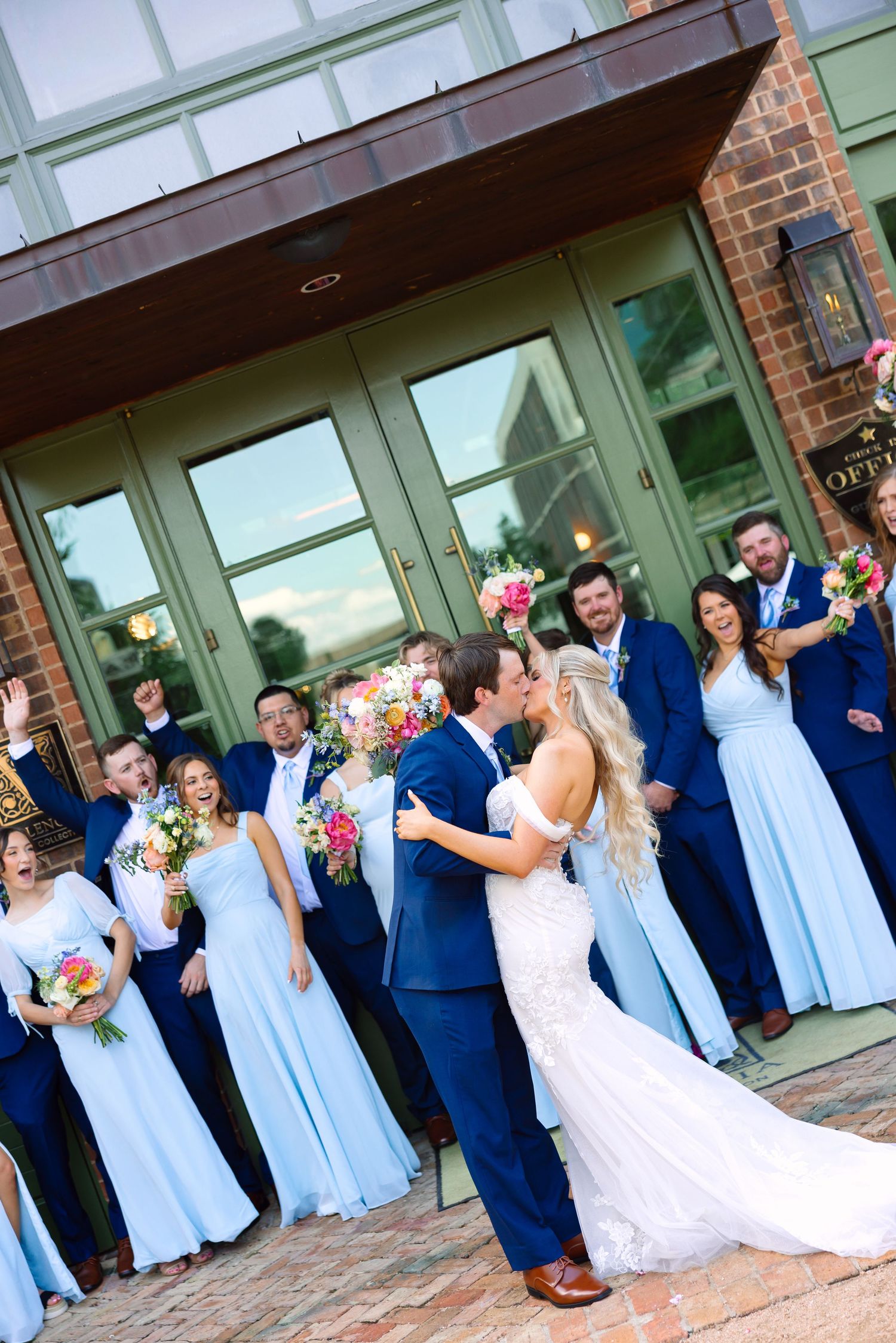 Wedding party in light blue dresses and navy suits cheering as bride and groom kiss.