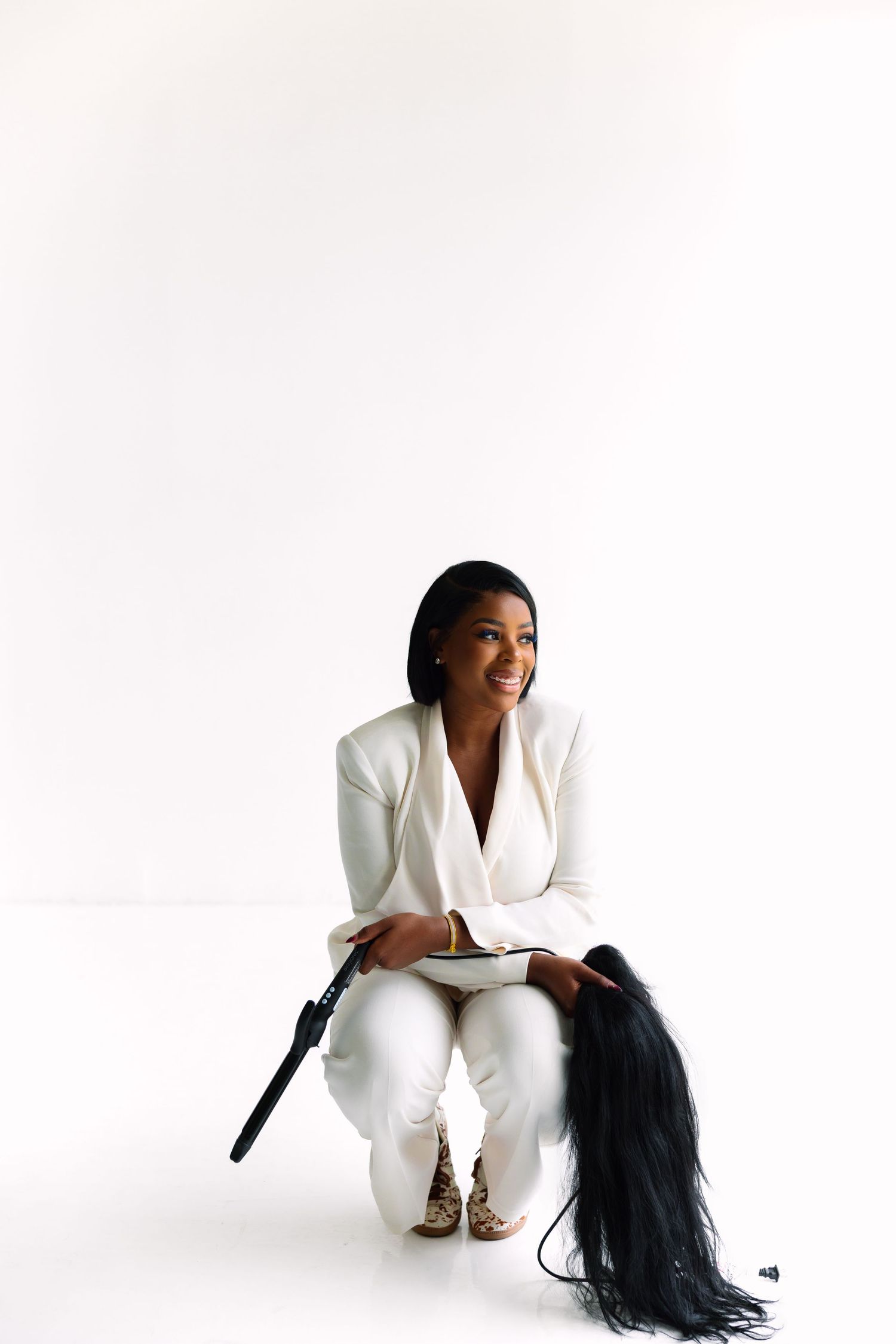 Woman in white suit sitting and smiling, holding a hair straightener with long black hair extensions.