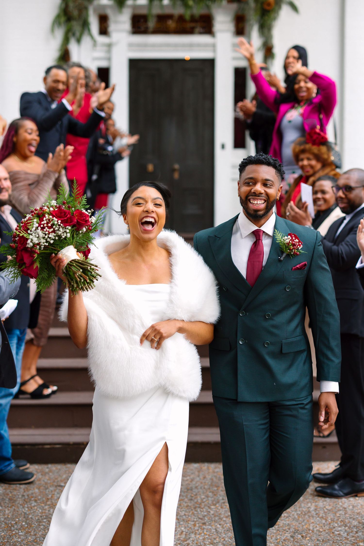 Joyful newlyweds exit ceremony as guests cheer, bride holds red bouquet wearing white gown with fur wrap, groom in green suit.