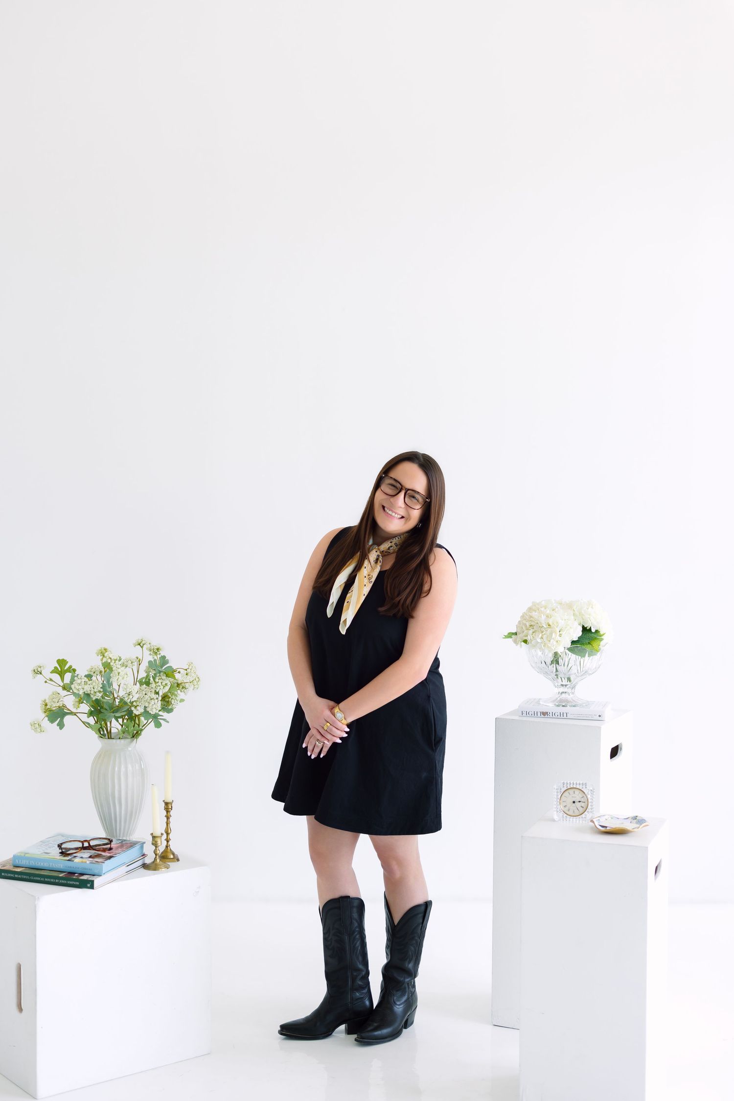 Woman in black dress and cowboy boots poses in white studio with floral arrangements on white pedestals.