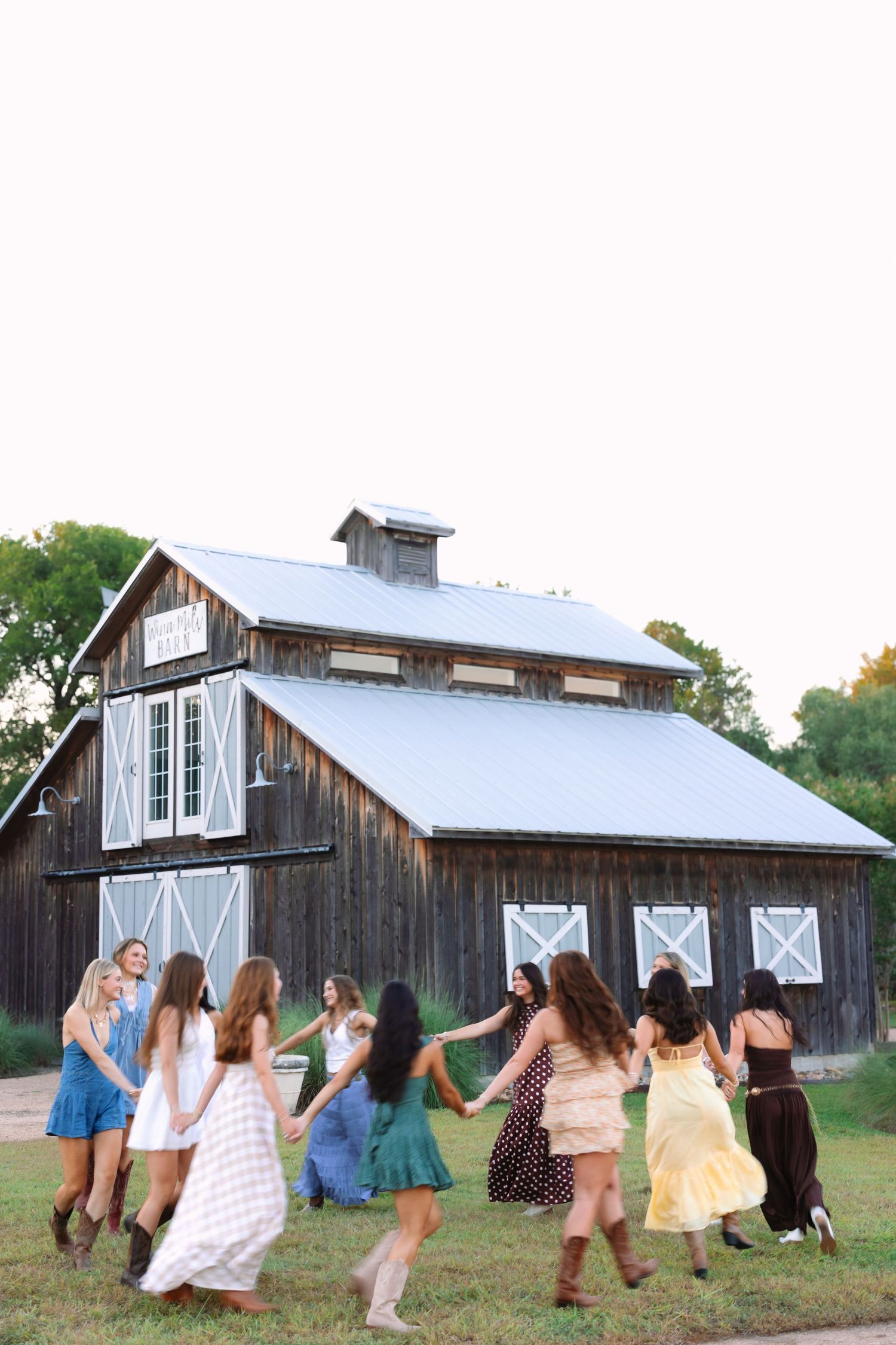 Friends dancing together in summer dresses near a wooden barn at sunset.