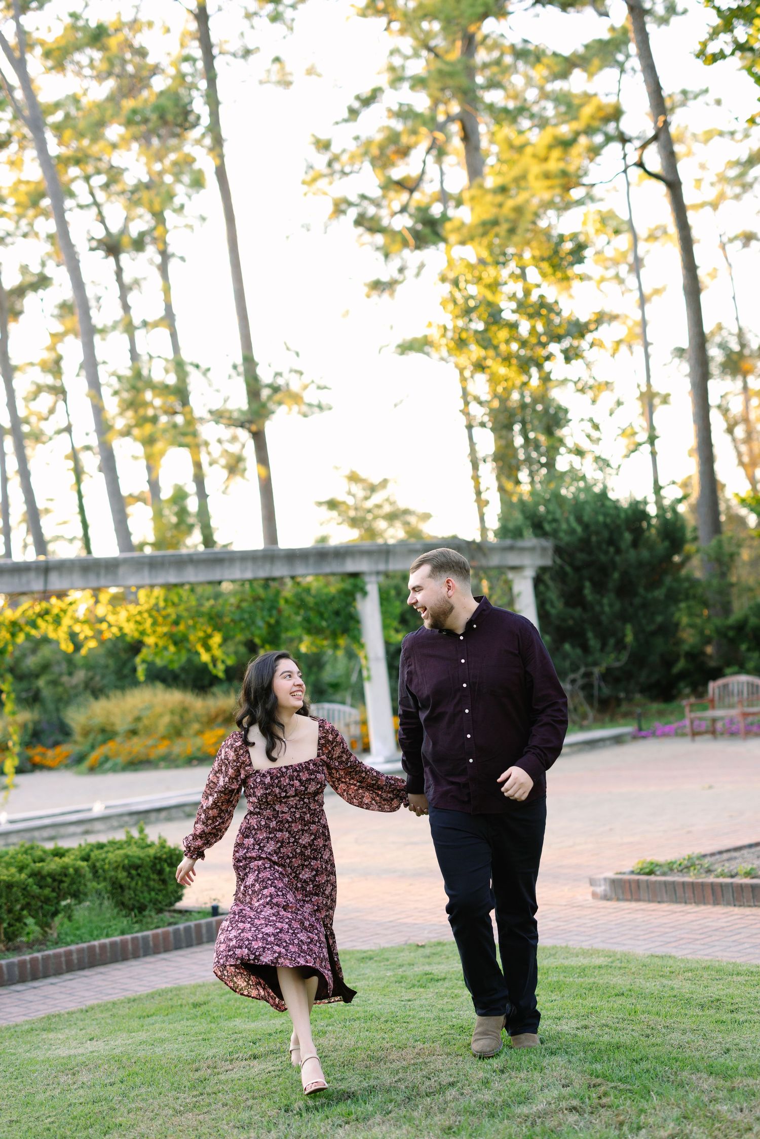 Couple holding hands and smiling while walking through a lush garden park, woman in floral dress and man in dark outfit.