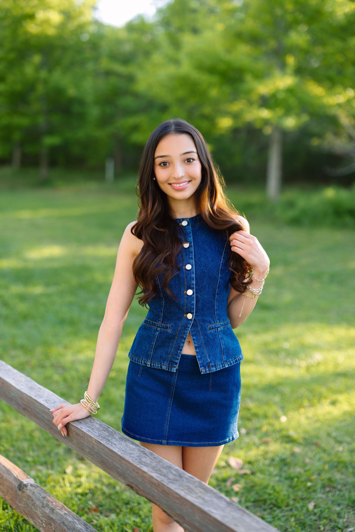 Young woman in denim skirt set smiling outdoors by wooden fence with lush green trees in background.