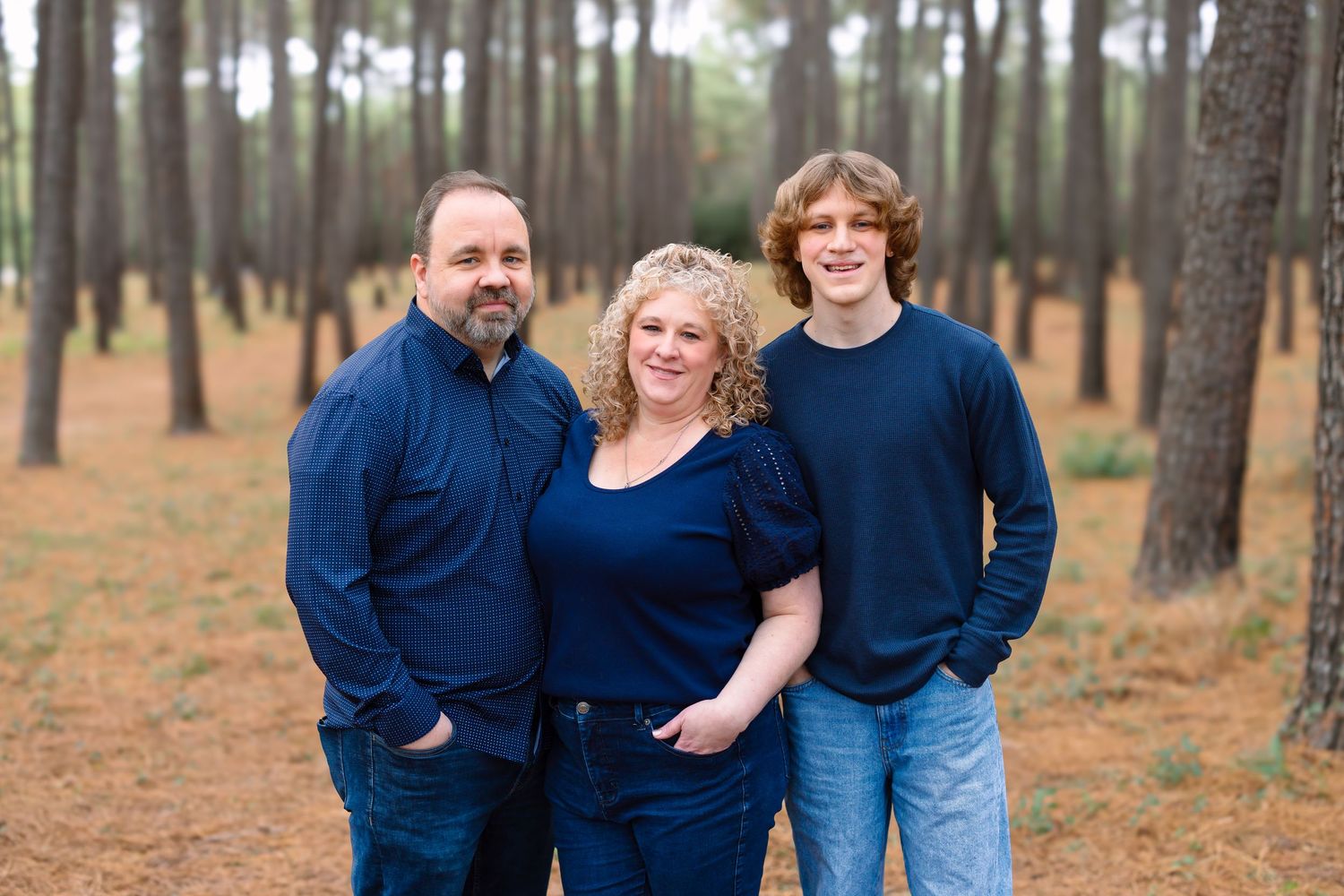 Family of three posing among pine trees in coordinating navy outfits for an outdoor portrait session.