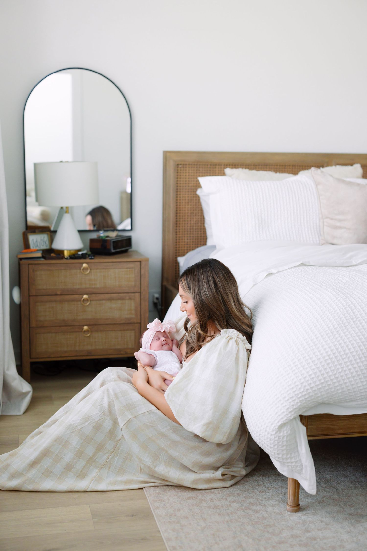 Mother sitting on bedroom floor cradling newborn baby, beside rattan bed and wooden nightstand.