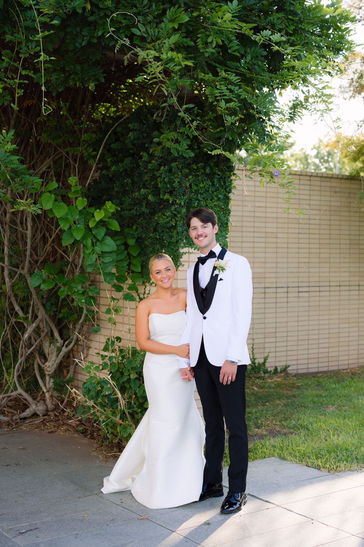 Bride in strapless white gown and groom in white tuxedo jacket pose together outdoors by a brick wall with lush greenery.