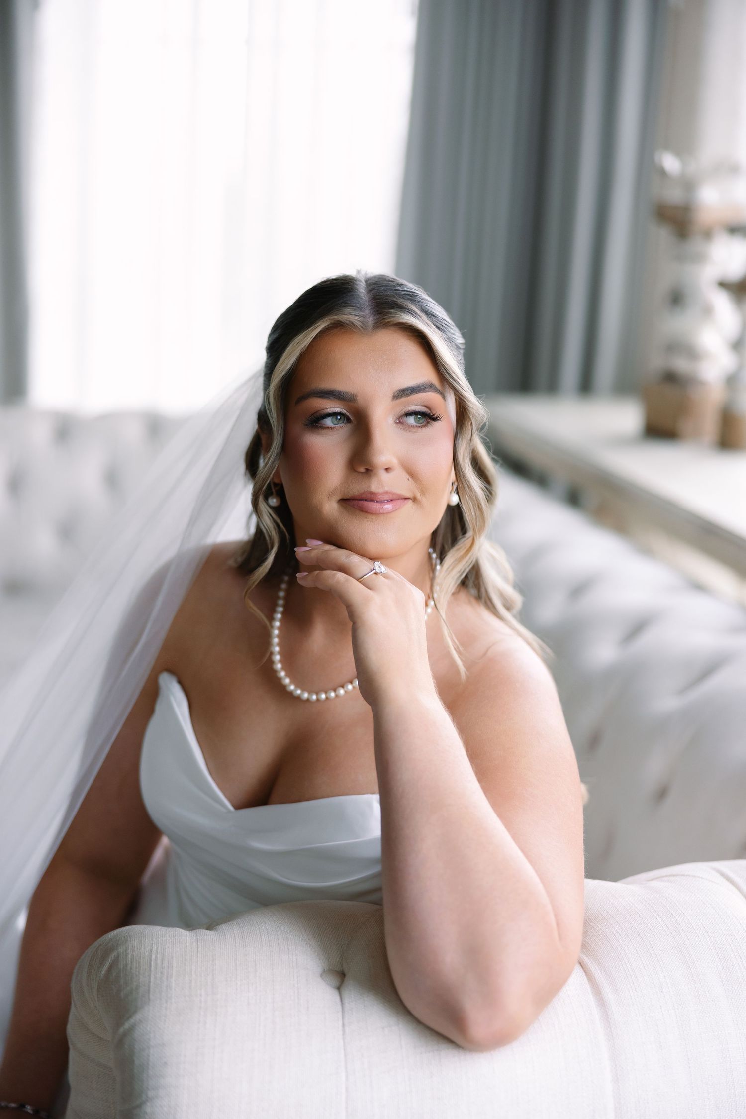 Bride in strapless white gown and veil sits on tufted sofa wearing pearl necklace, gazing thoughtfully.